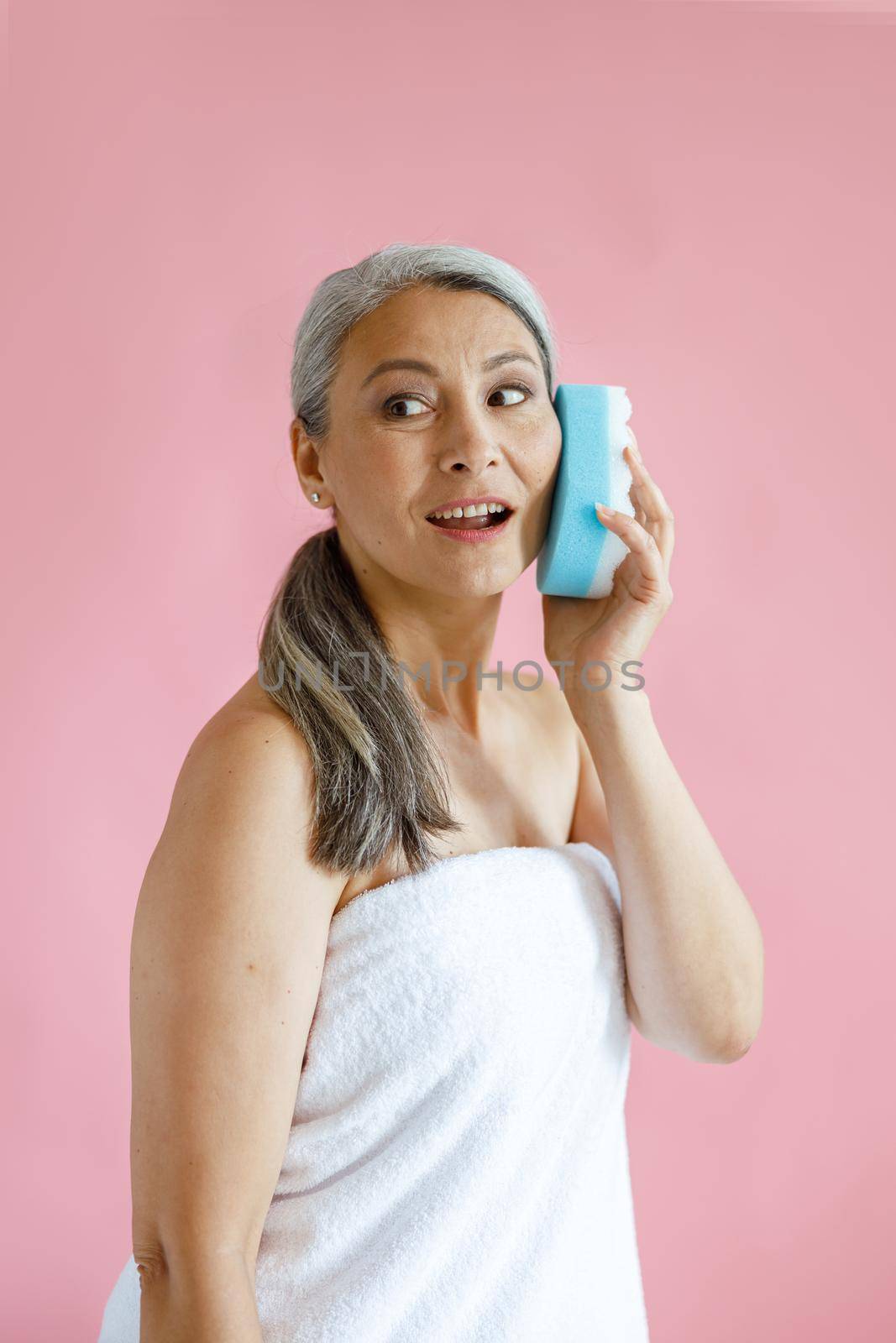 Beautiful hoary haired Asian woman with towel uses bath sponge as mobile phone on pink background in studio. Mature beauty lifestyle