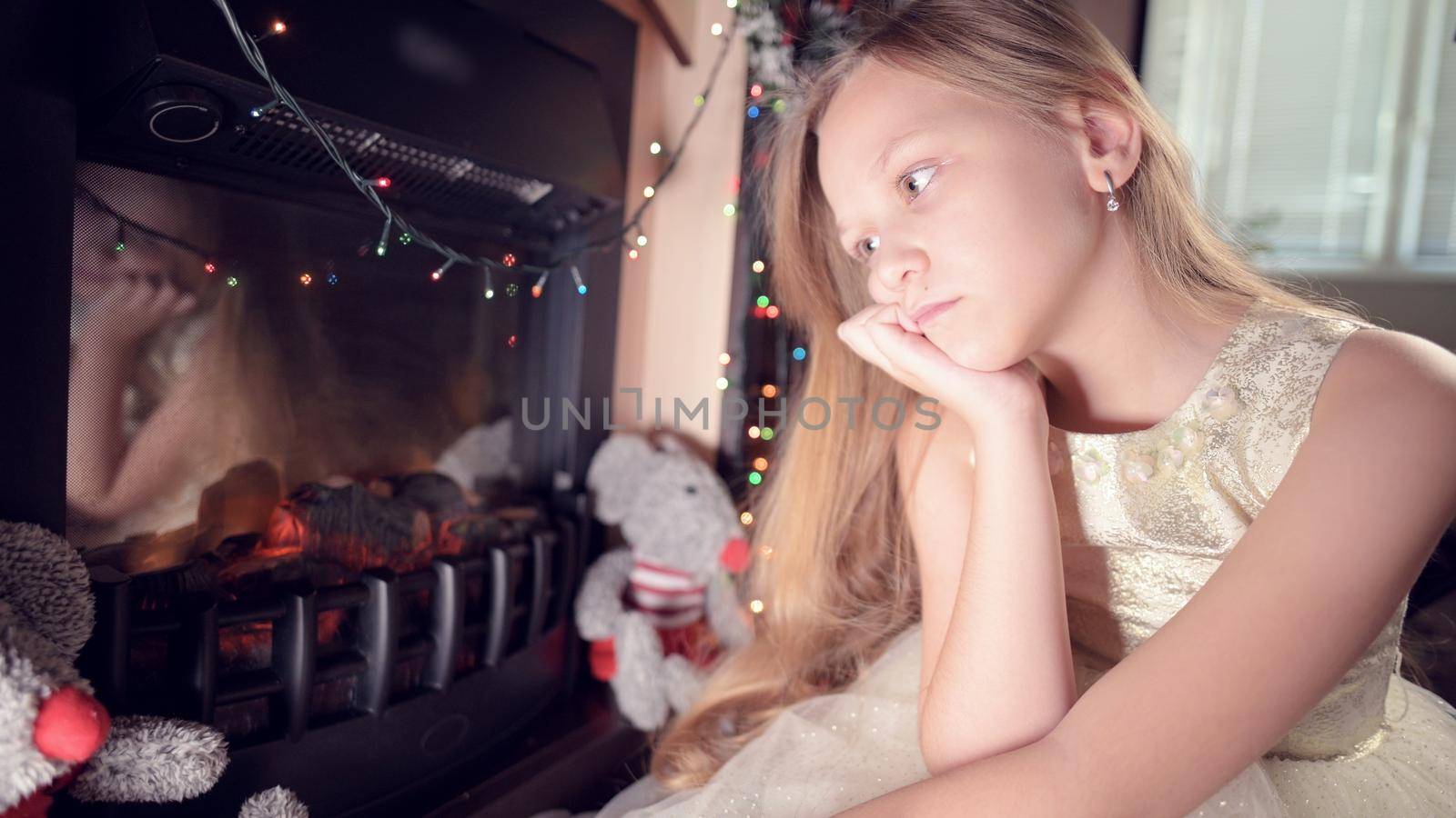 A little sad disappointed girl sits by an artificial fireplace and is sad next to soft toys. The concept of spoiled Christmas holiday by yanik88