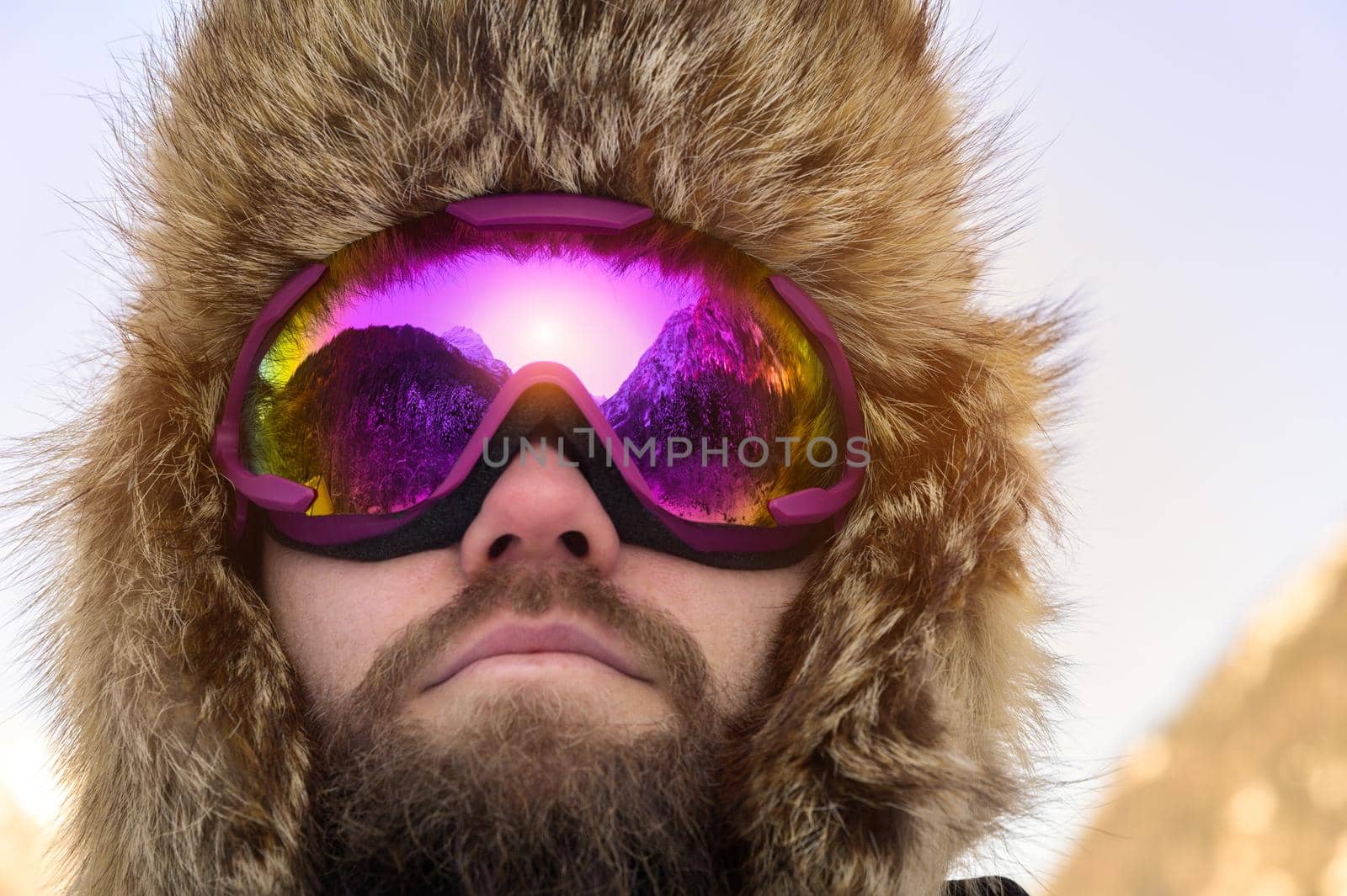 Close-up portrait of a bearded happy snowboarder skier in a ski mask with goggles and a fur big old-school hat. on a background of a winter snowy mountains.