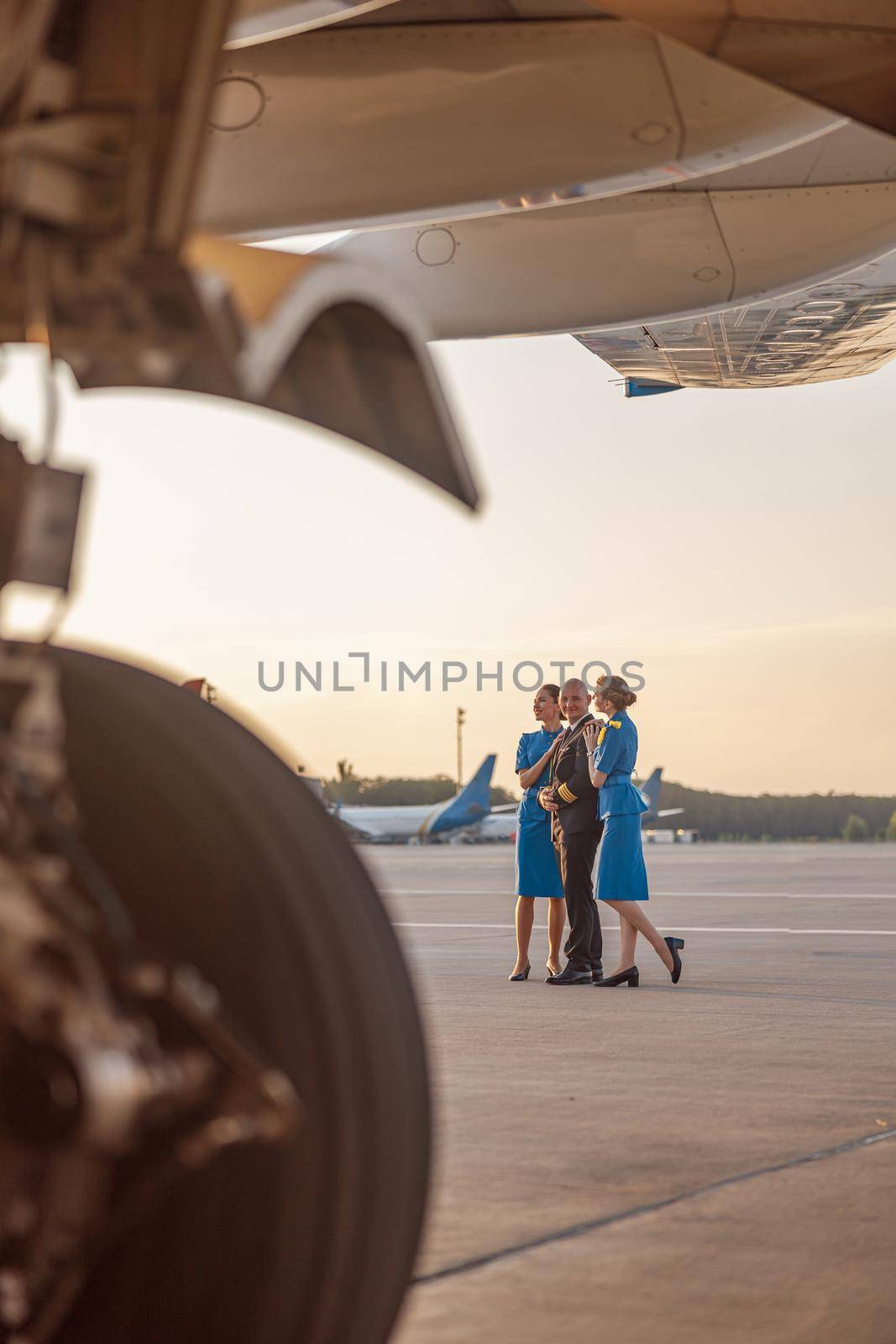 Full length shot of male pilot posing for photoshoot together with two air stewardesses in blue uniform, standing in an airport terminal at sunset by Yaroslav_astakhov