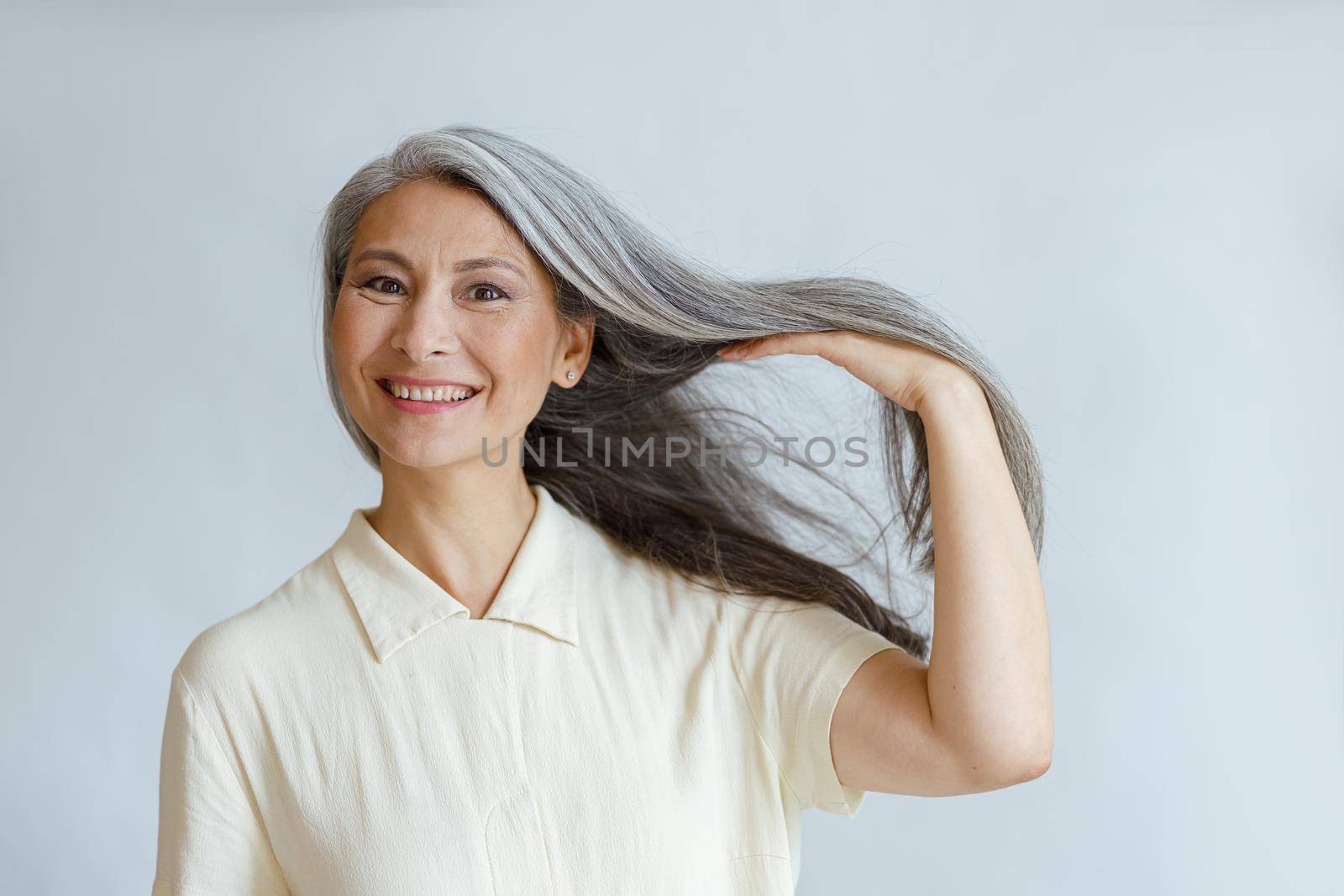 Happy middle aged Asian lady shows natural silver hair posing on light background by Yaroslav_astakhov