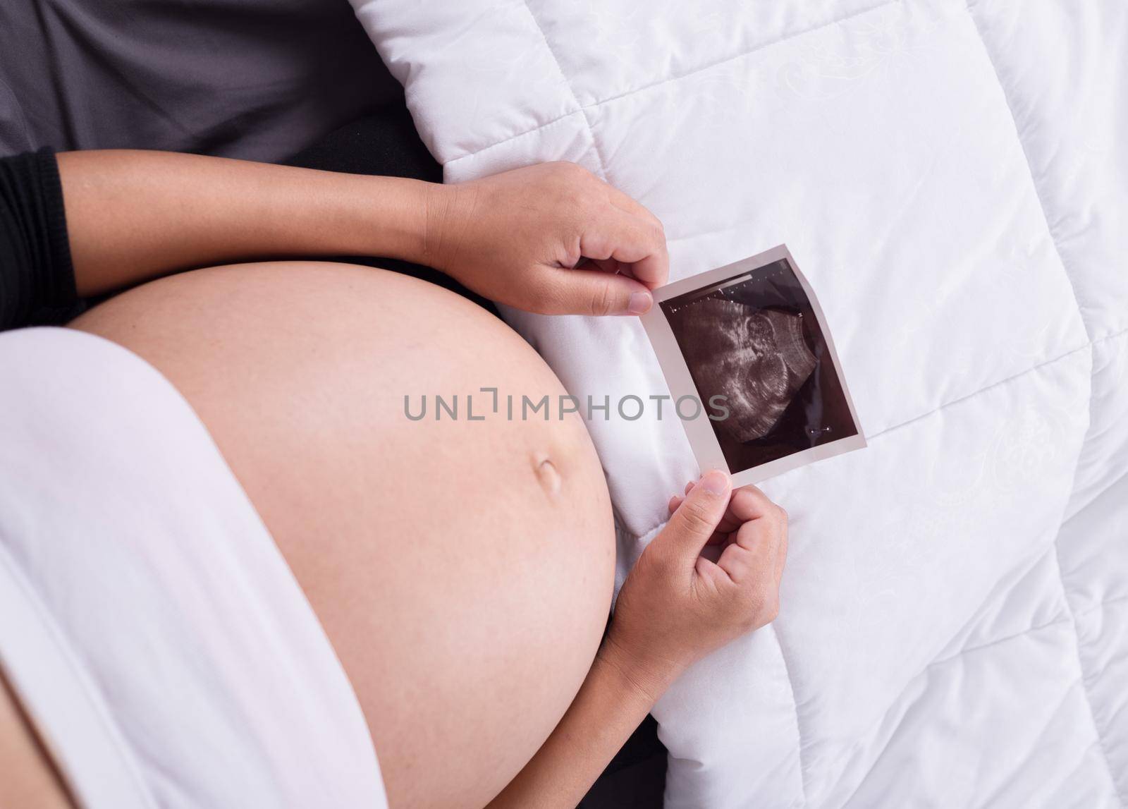 pregnant woman with ultrasound image of baby sitting on bed in bedroom at home by geargodz
