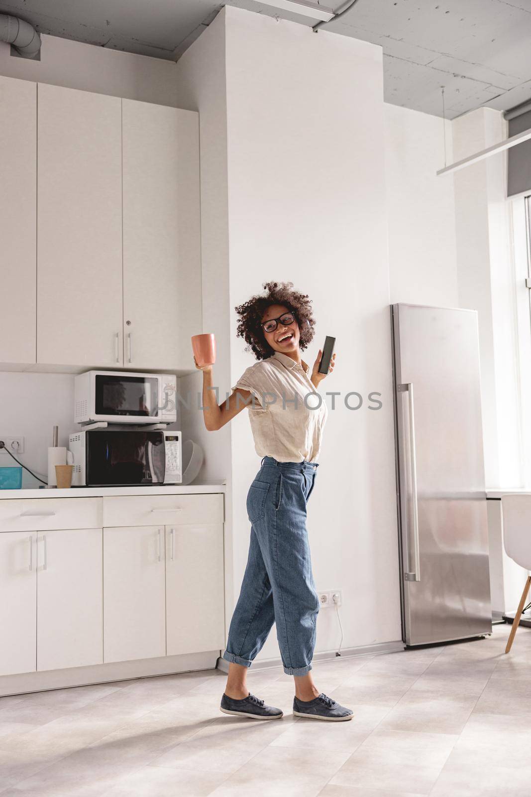 Full-length photo of smiling pretty lady holding cup while dancing on the kitchen and using headphones