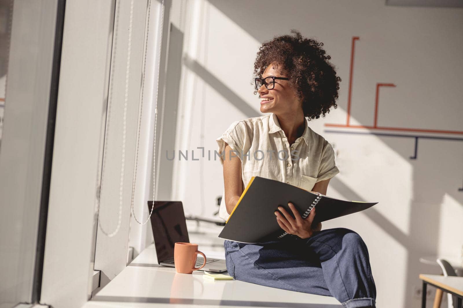 Happy Afro American female worker sitting on the table while holding notebook and looking out the window