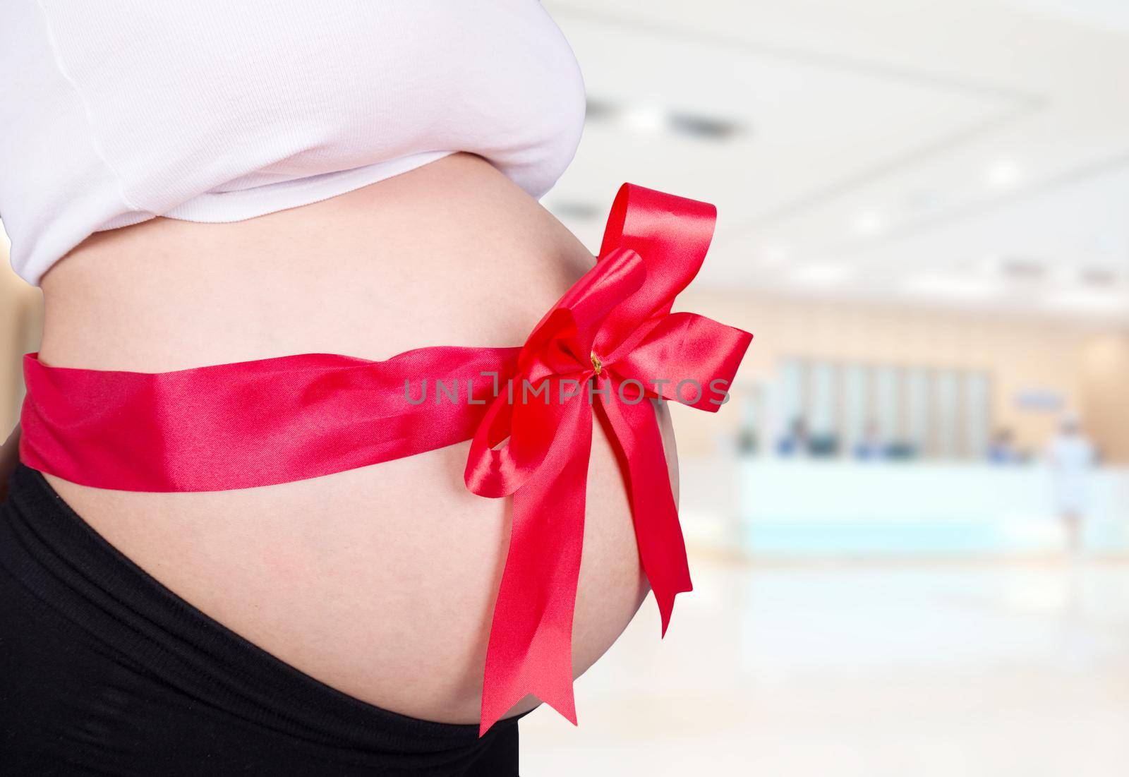 close up pregnant woman with red ribbon gift on belly in hospital background by geargodz