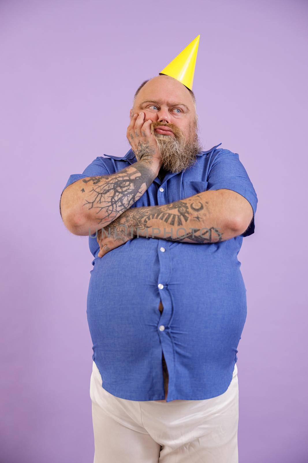 Bored middle aged man with overweight in tight blue shirt and party hat leans onto palm on purple background in studio