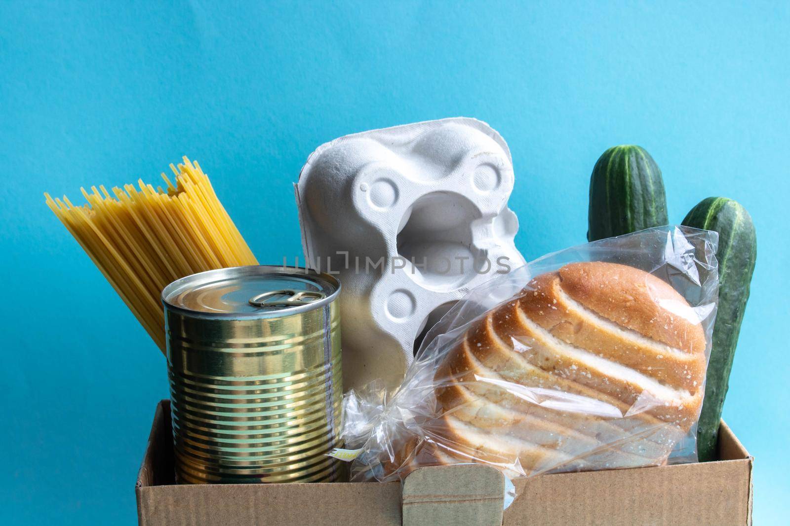 Home delivery of products.Helping people during a pandemic.A box of food stands on a blue background.Spaghetti, cucumbers, tushonka in a jar, bread and a container with eggs are in the box by lapushka62