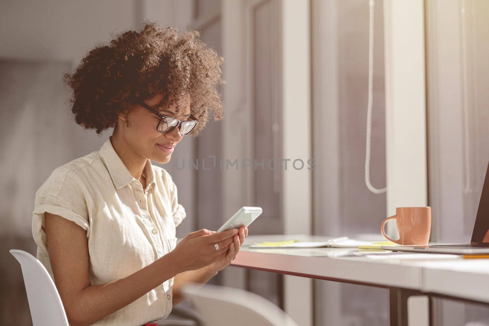 Pretty multiethnic lady wearing glasses and holding smartphone and looking at screen in the office