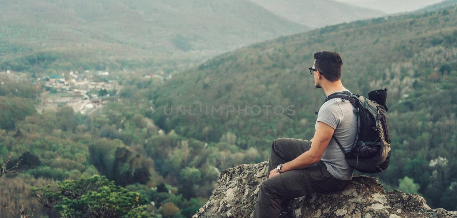 Traveler young man with backpack sitting on top of rock and looking into the distance