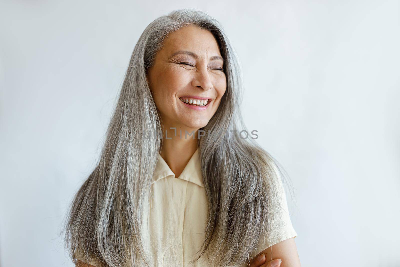 Pretty laughing Asian lady with natural silver hair poses for camera on light grey background in studio. Mature beauty lifestyle