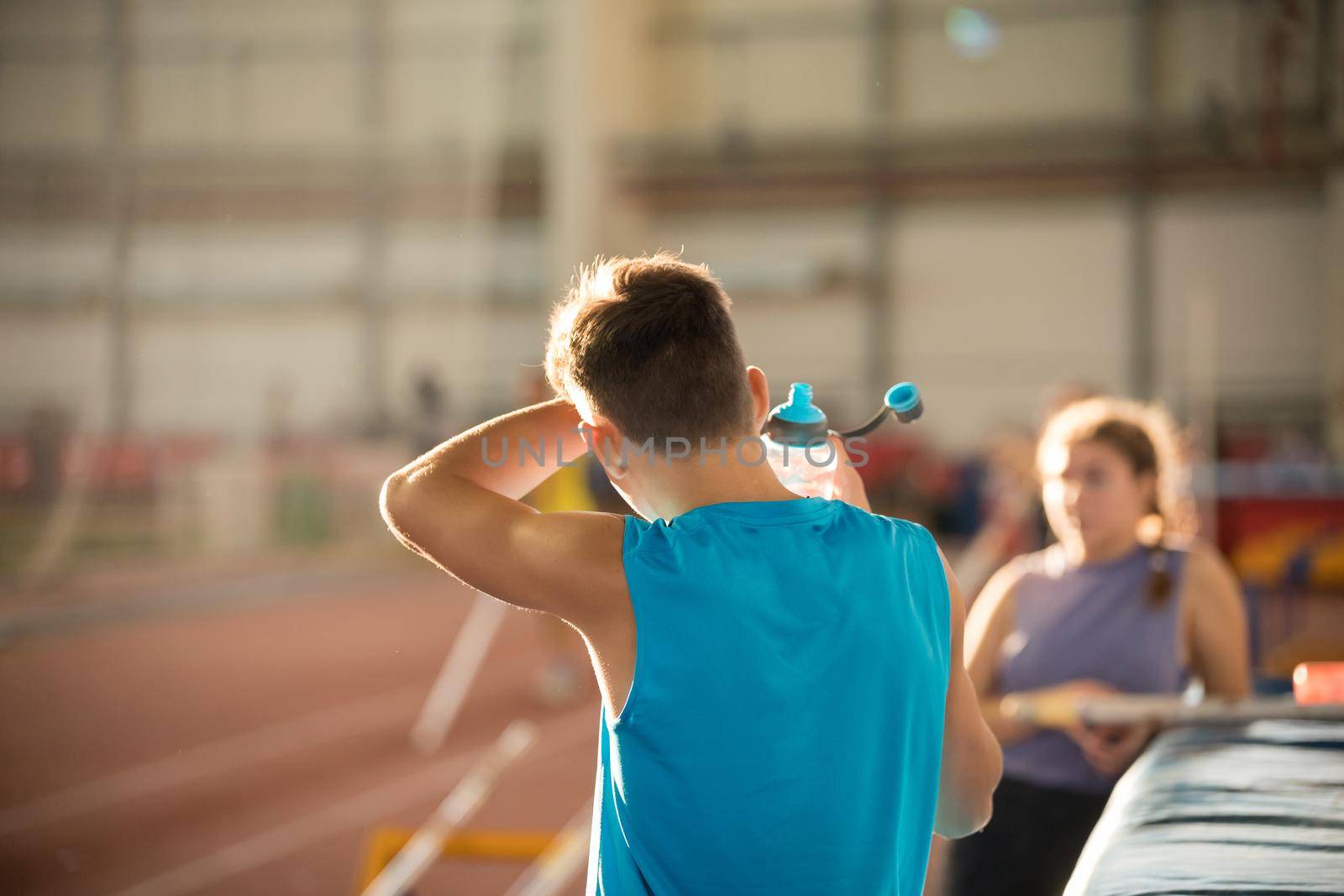 Young man in blue shirt standing on the sports stadium holding a water bottle - wiping out the sweat from his forehead. Mid shot