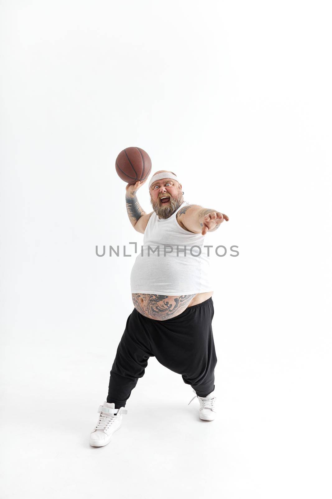 bearded fat caucasian tattoed man with basketball ball in white background. He is funny and happy