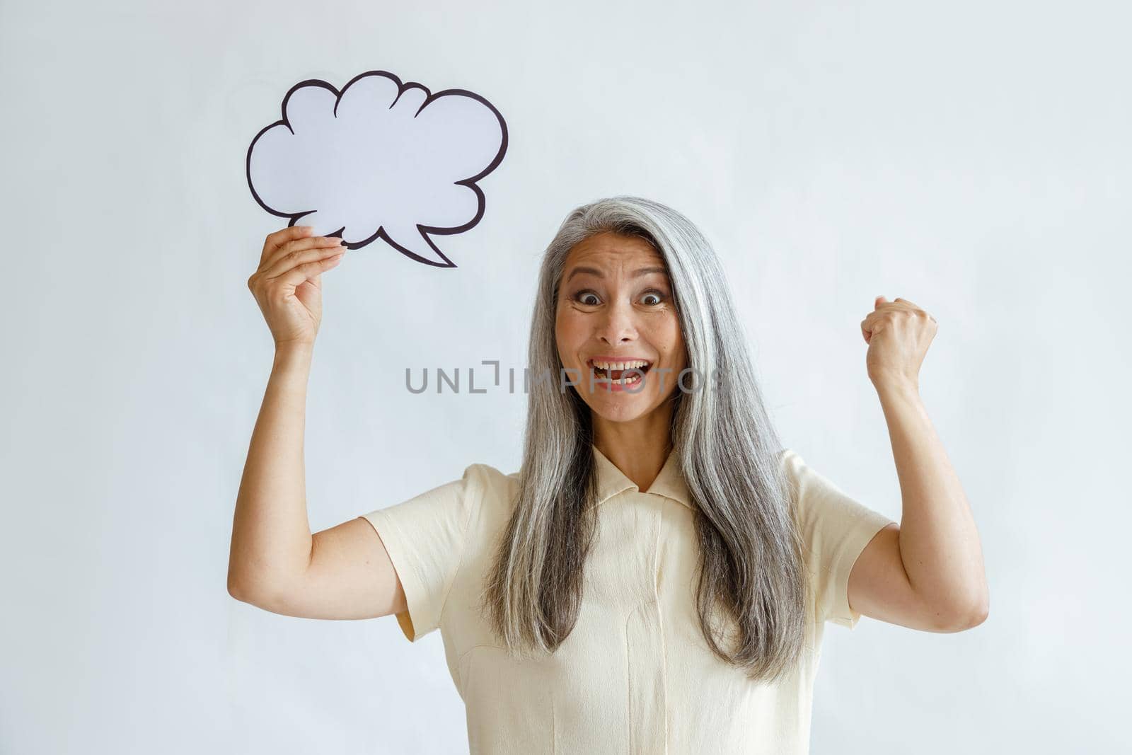 Excited mature Asian female model with hoary hair holds blank text bubble posing on light background in sudio. Mockup for design