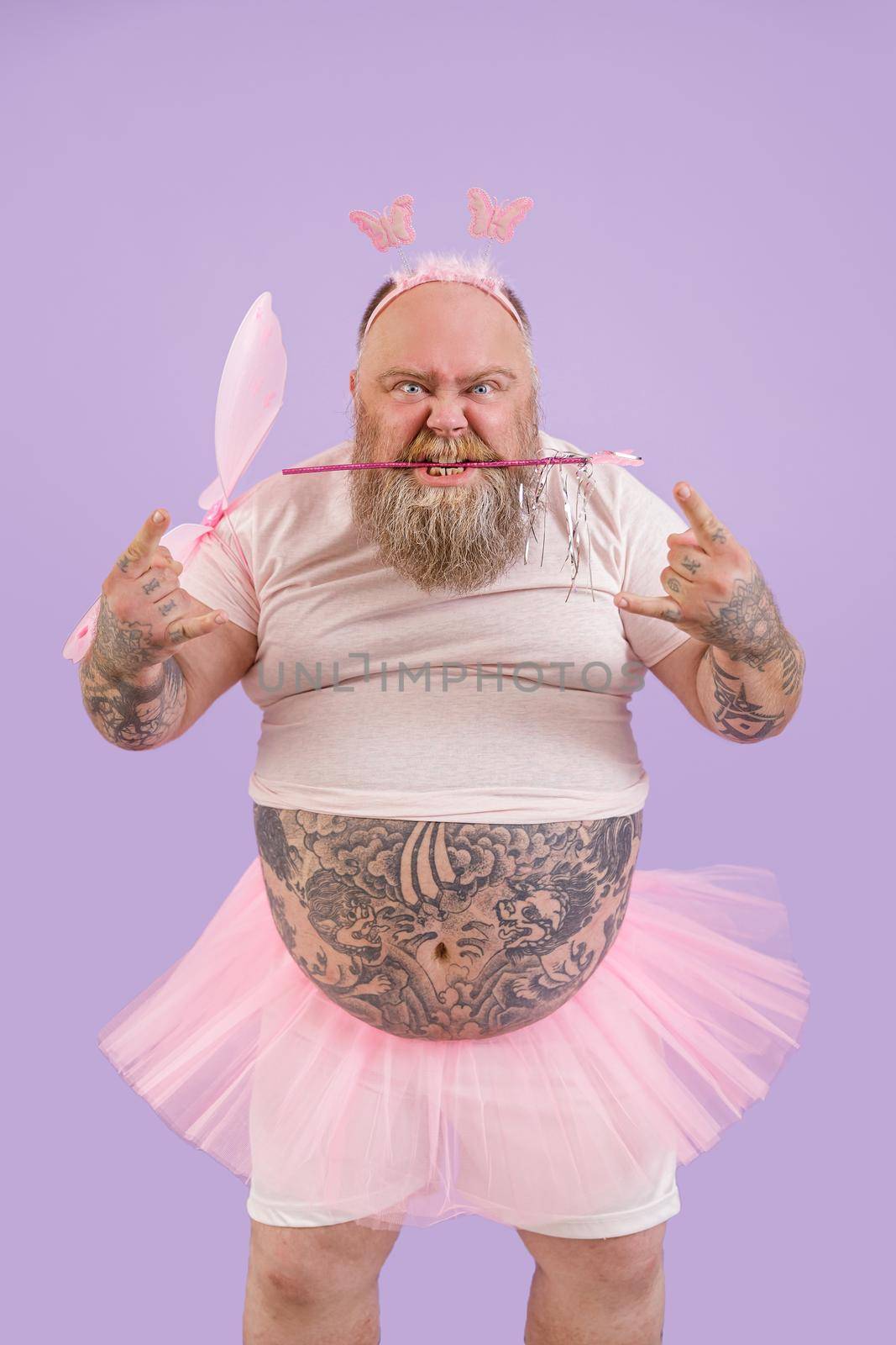 Brutal person with overweight and tattoos in fairy suit with wings holds magic stick in teeth and shows horns gesture on purple background in studio