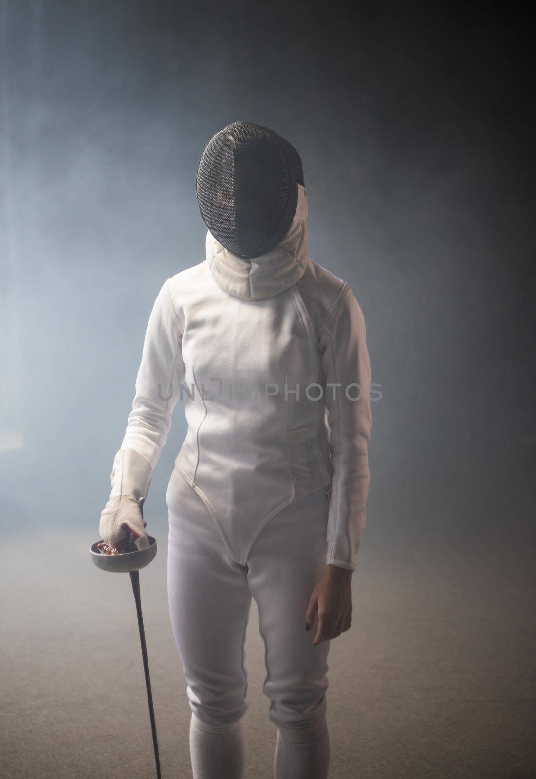 A young woman fencer in protective helmet standing with a sword down. Mid shot