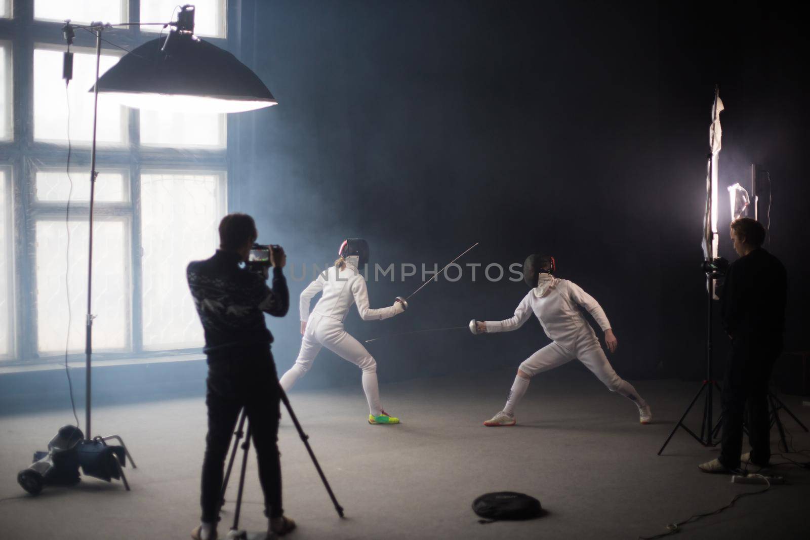 A fencing training in the studio - two women in protective costumes having a duel - a cameraman standing near the tripod and shooting the duel. Mid shot