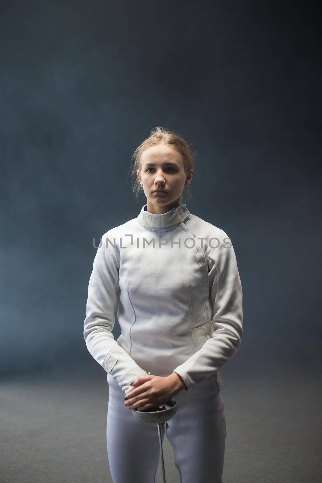A young woman fencer with her hair in a bun standing with a sword down. Mid shot