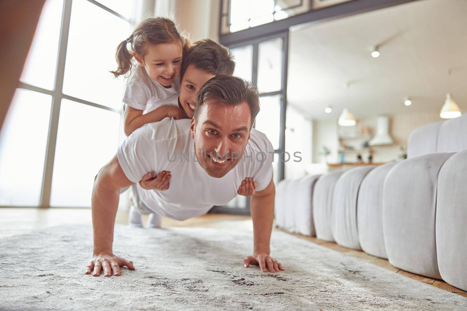 Portrait of smiling father exercising while holding daughter and son on his back during workout indoors
