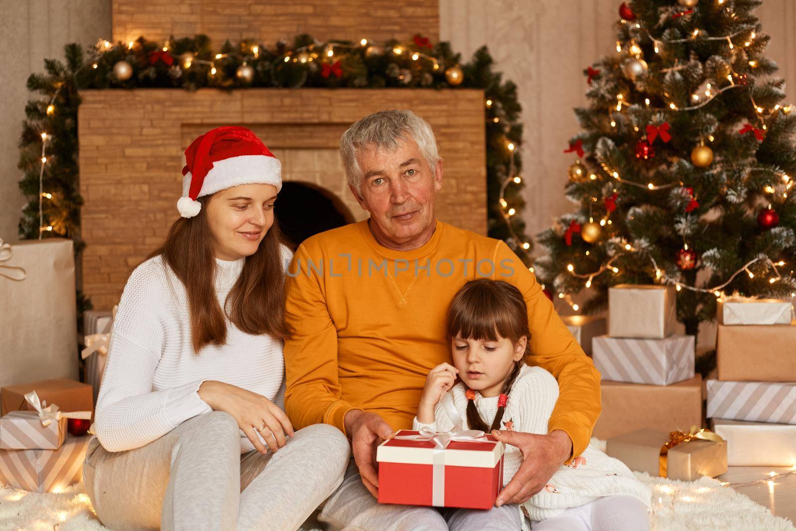 Indoor shot of happy family giving Christmas present to little cute dark haired girl in red wrapped gift box, posing in decorated living room near xmas tree and fireplace. by sementsovalesia