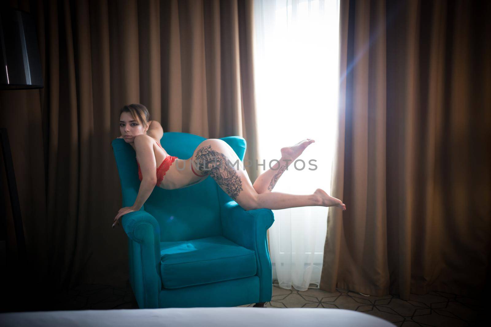 Young woman with tattoos in a sexy red underwear graceful standing in the chair on her knees. Looking in the camera