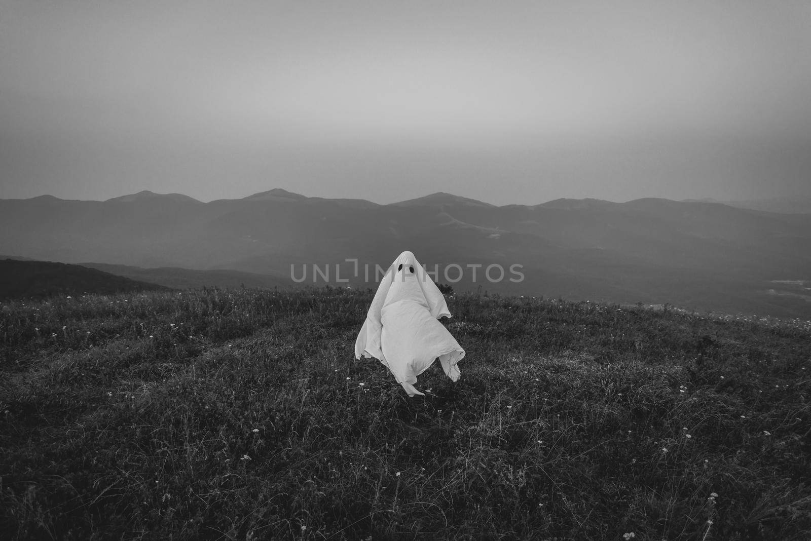 White ghost flying on meadow outdoor, monochrome image. Man in costume of Halloween ghost