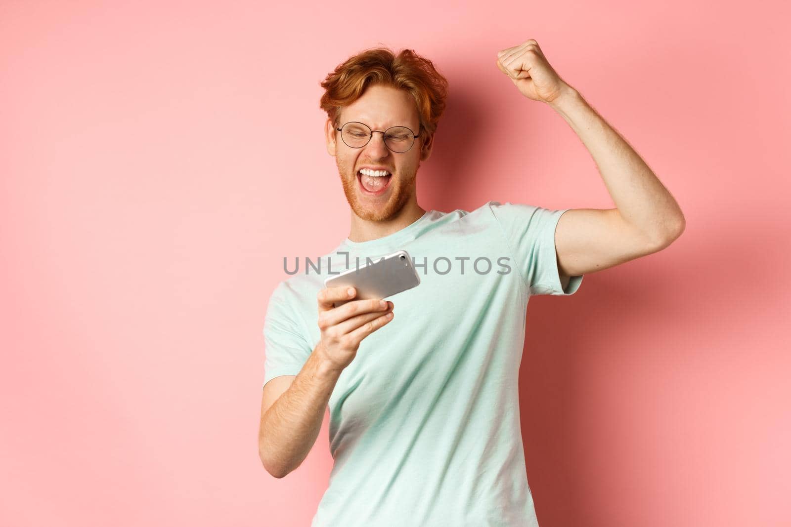Happy redhead man winning in mobile video game, raising hand up and shouting yes with joy, celebrating victory, looking at smartphone, standing over pink background by Benzoix