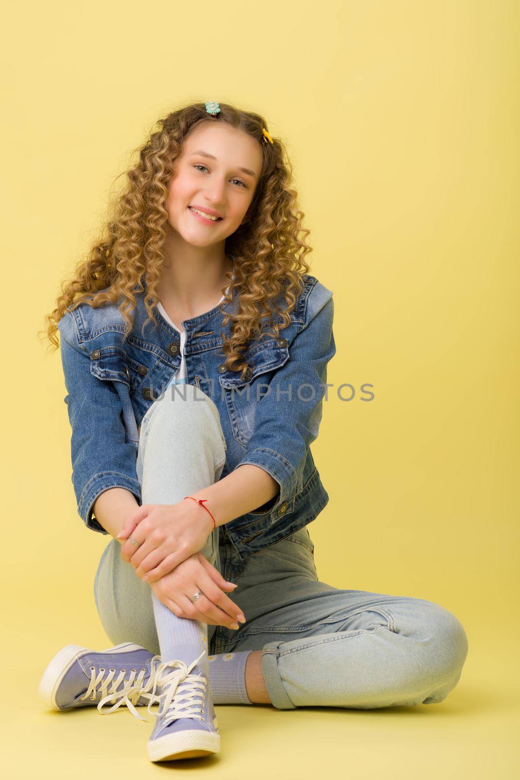 Happy girl sitting on floor hugging her knee. Carefree beautiful blonde girl with curly hairstyle wearing stylish denim jacket and jeans sitting on floor on yellow background