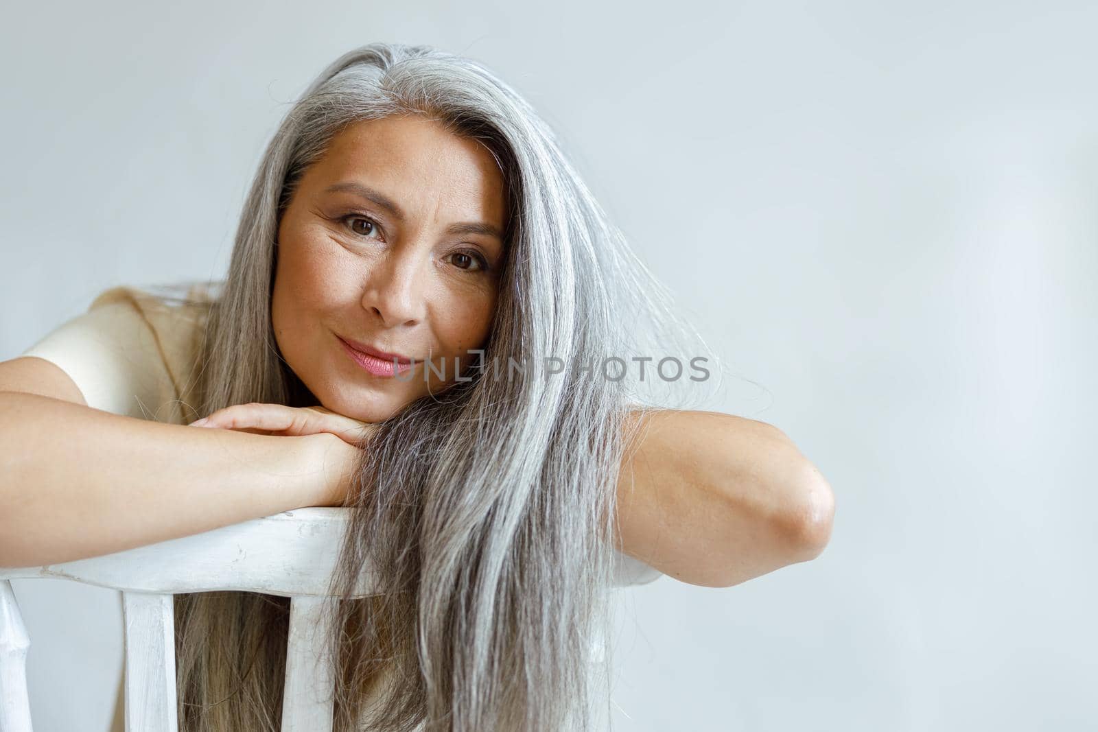 Cheerful middle aged Asian lady with flowing hoary hair sits backwards on chair on light background in studio. Mature beauty lifestyle