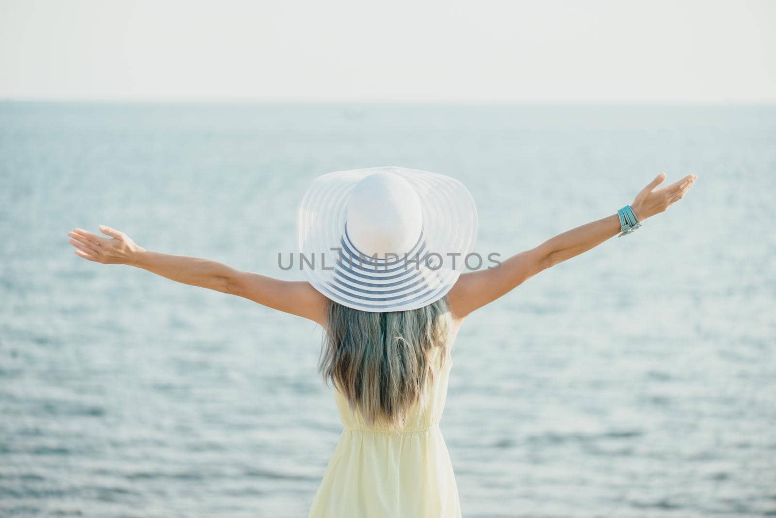 Happy young woman wearing in dress and hat with a wide brim standing with raised arms and enjoying view of sea, rear view