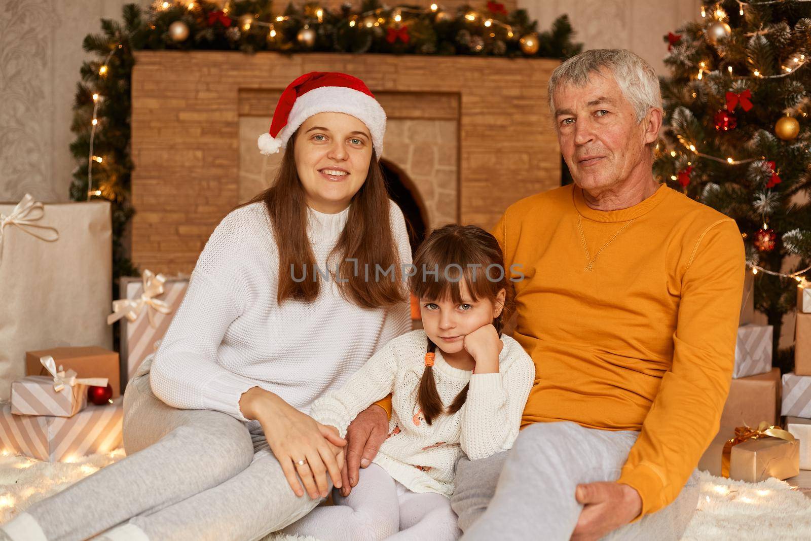 Portrait of happy family, mature man wearing orange sweater, his daughter and granddaughter sitting near Christmas tree at home, looking at camera with positive facial expressions. by sementsovalesia