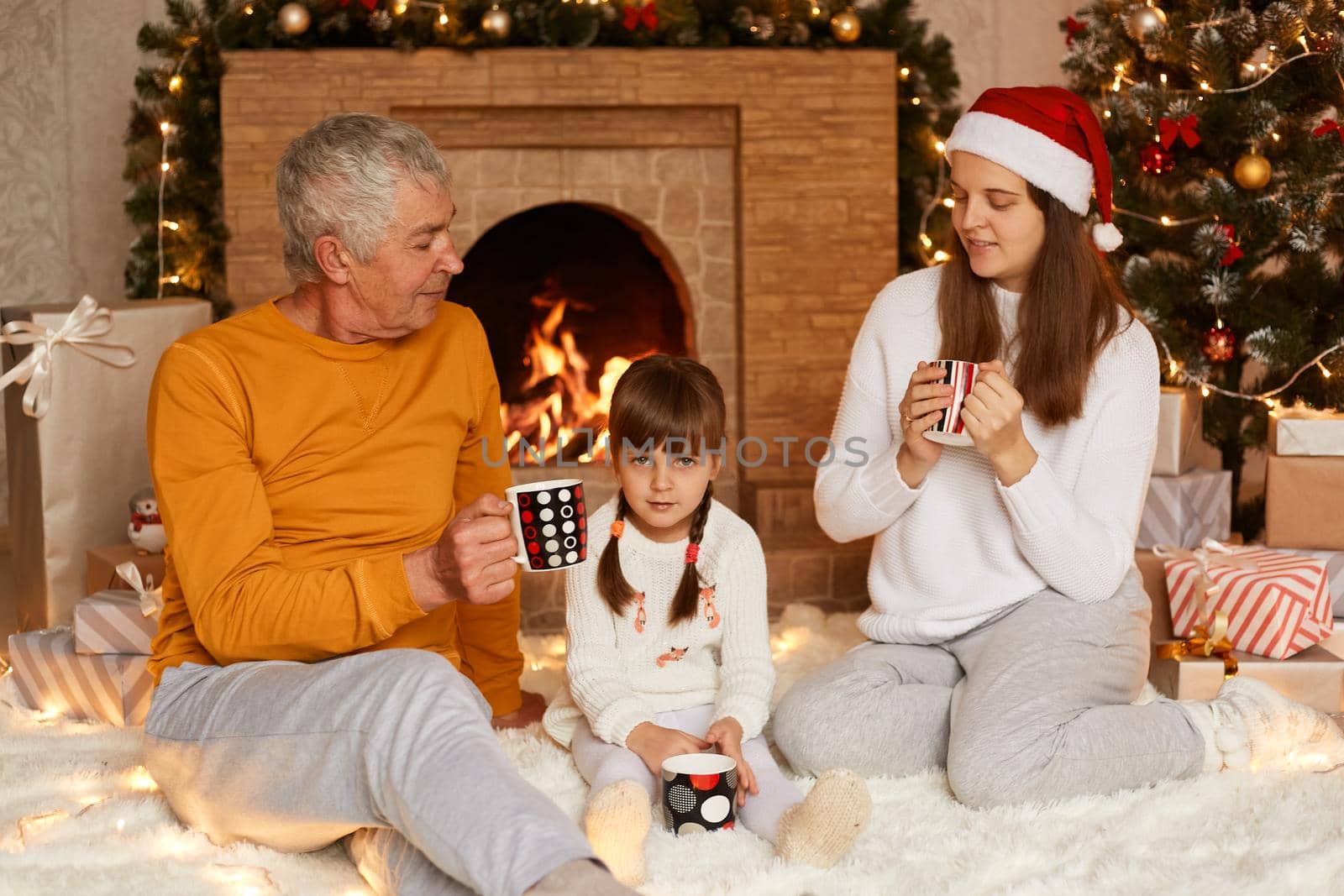Happy family senior man, woman and little girl sitting in festive room near fireplace and christmas tree and drinking hot tree, talking together during new year eve. by sementsovalesia