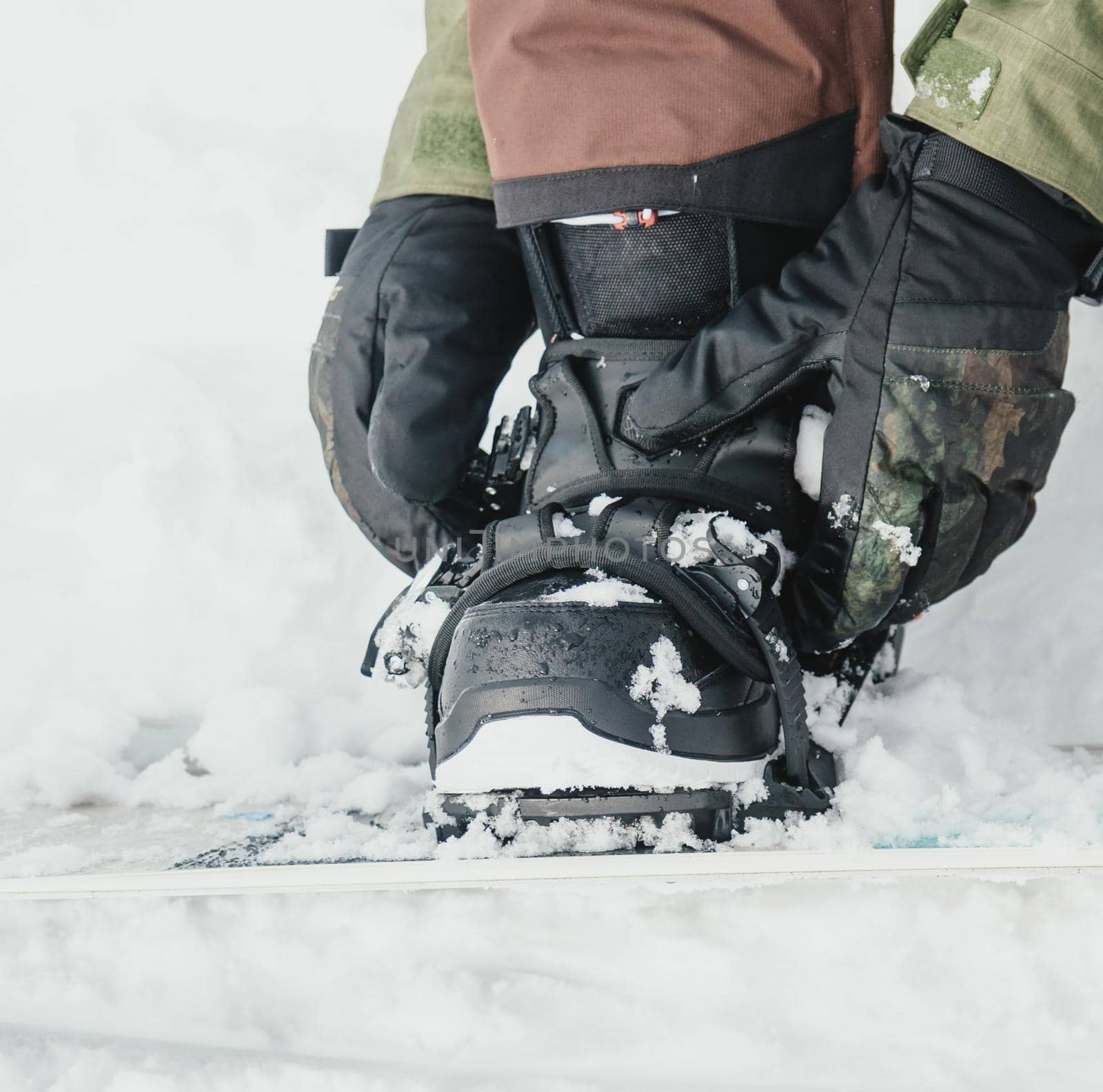 Young man putting on his snowboard and tightening the straps on background of snow in winter, close-up image