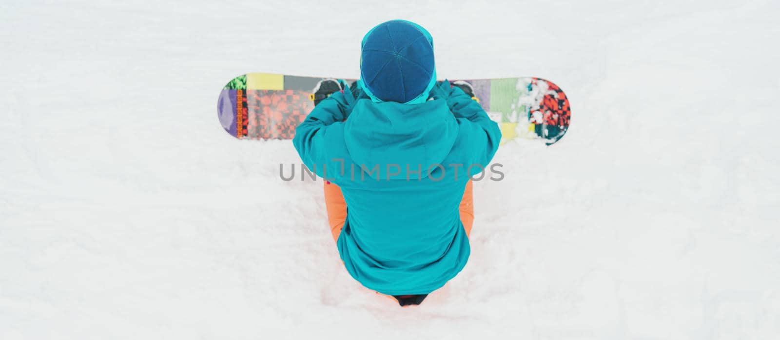 Unrecognizable young woman sitting with snowboard on snow, rear view