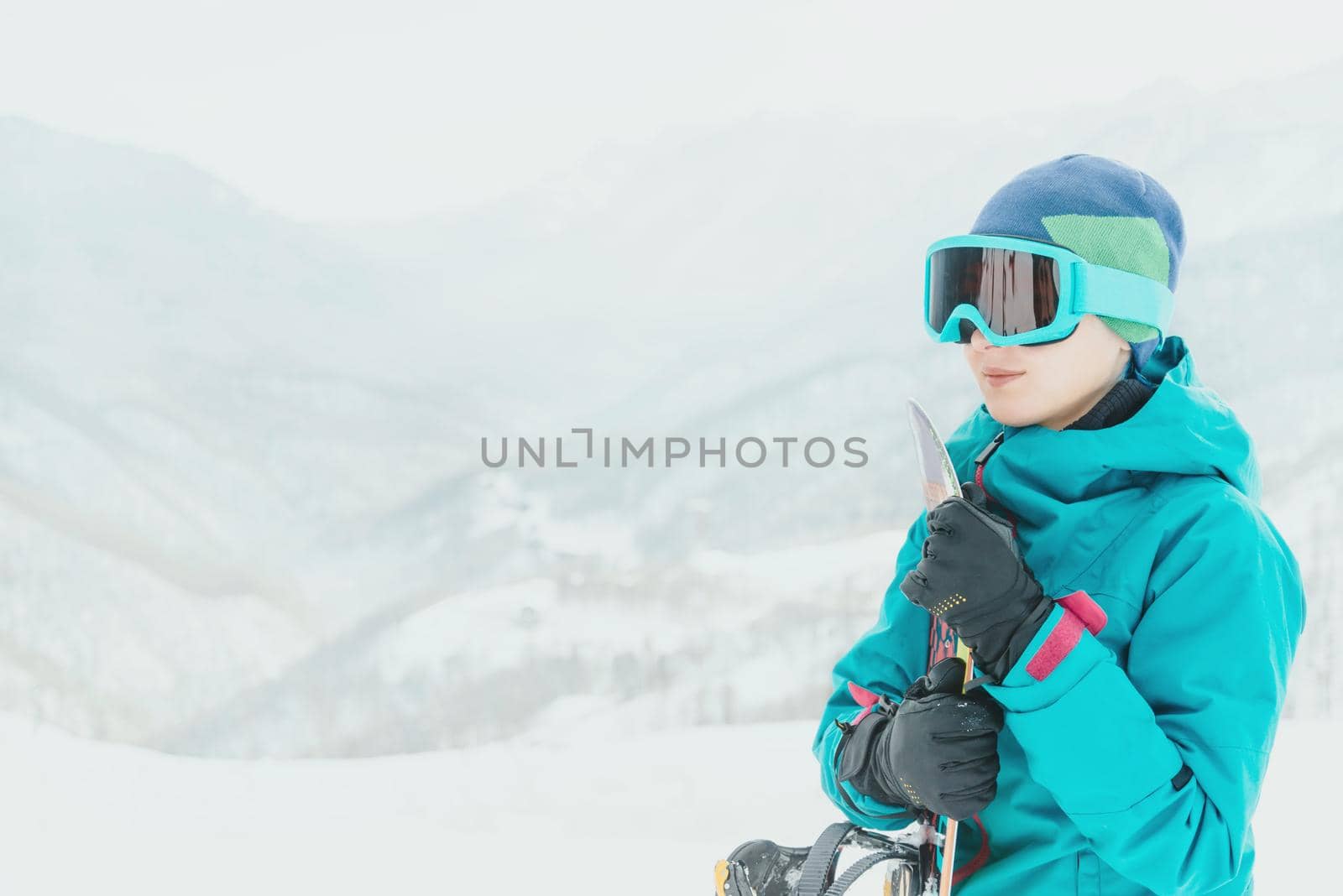 Smiling young woman in protective sunglasses standing with snowboard on background of snowy mountains. Space for text in left part of image.