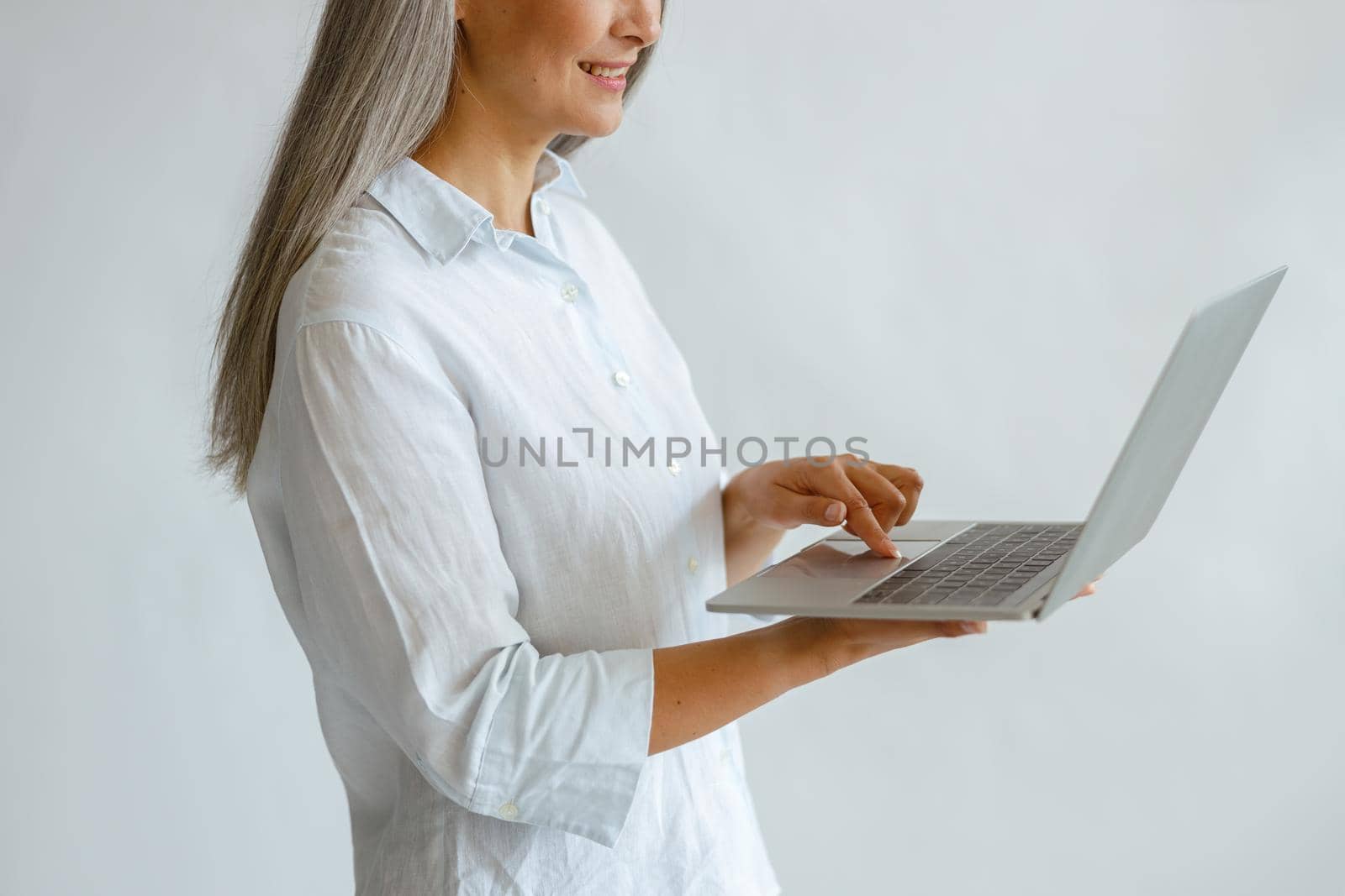 Hoary haired lady in white blouse uses laptop on light background in studio by Yaroslav_astakhov