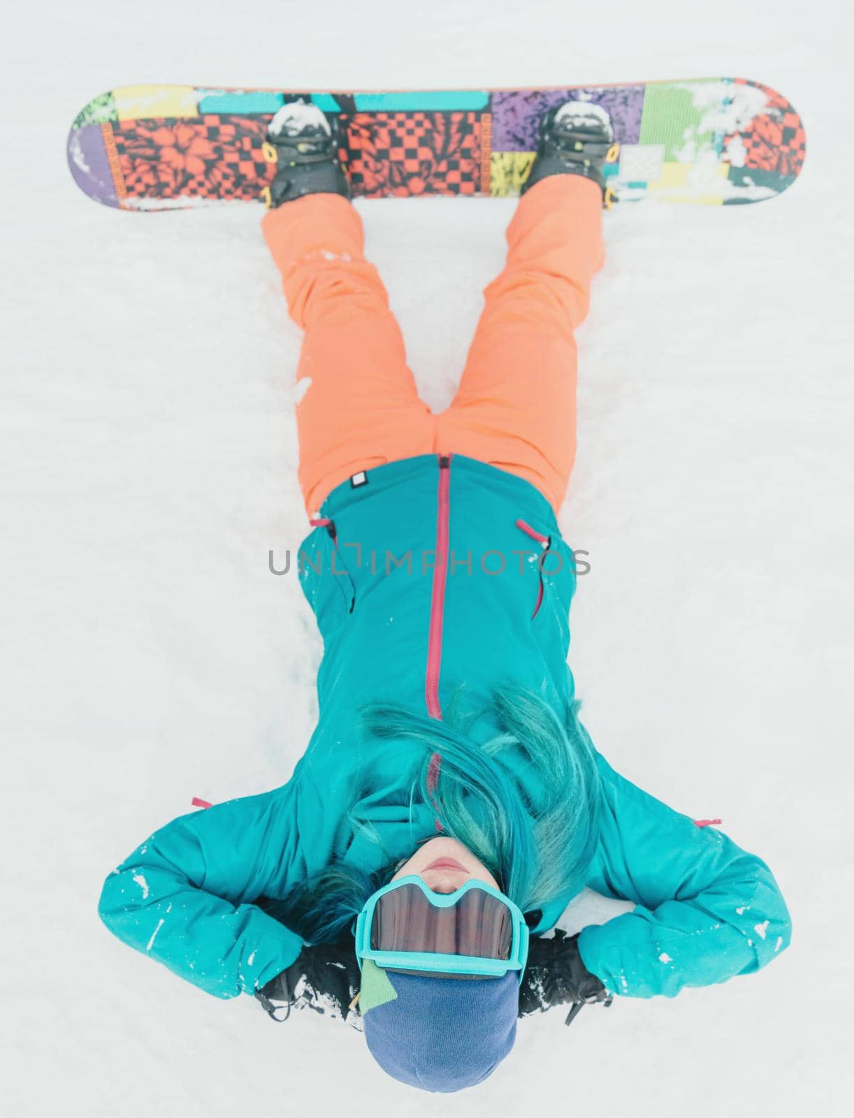 Young woman in protective sunglasses with snowboard lying on snow, top view