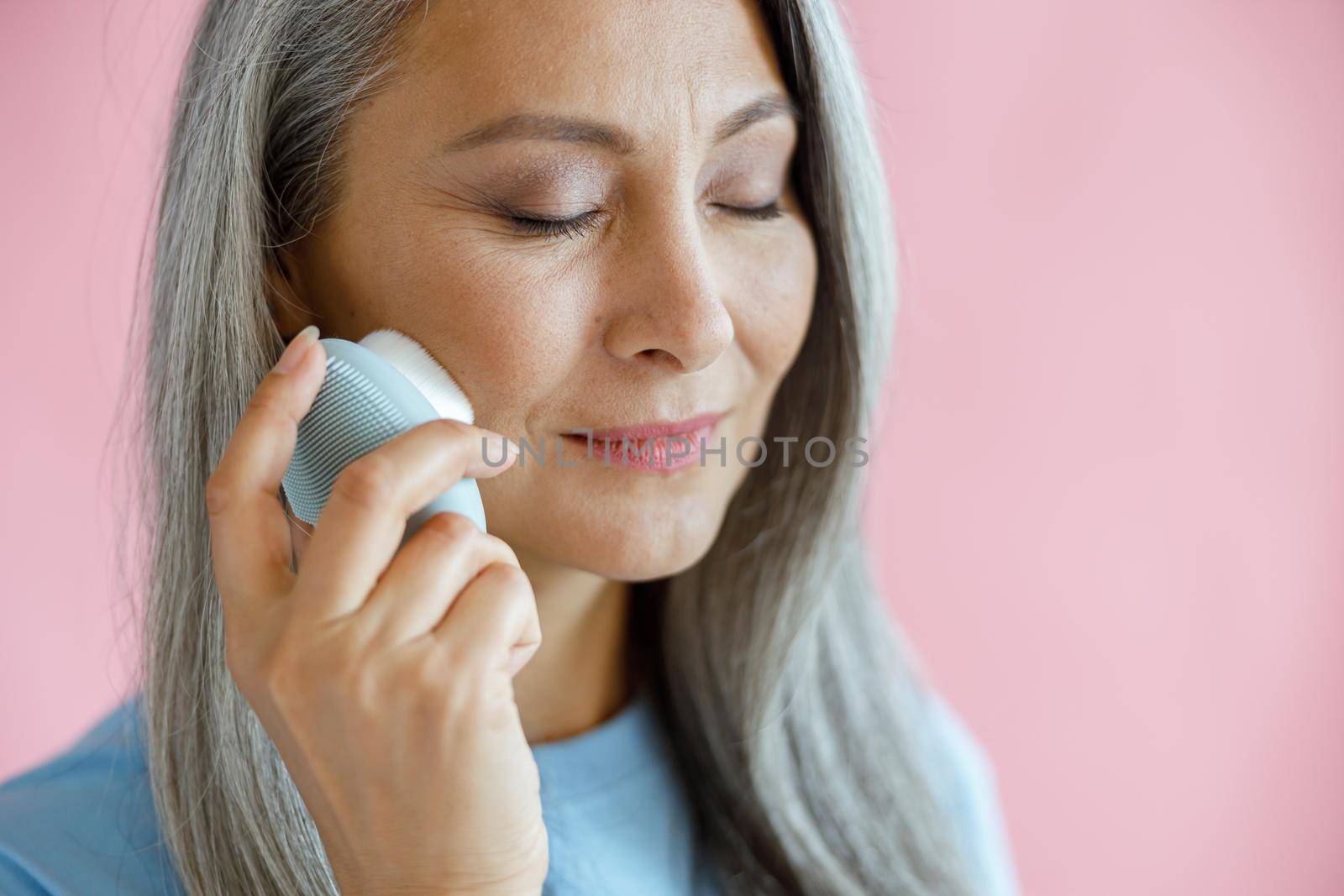 Beautiful silver haired Asian lady uses sonic facial cleansing brush standing on pink background in studio. Mature beauty lifestyle