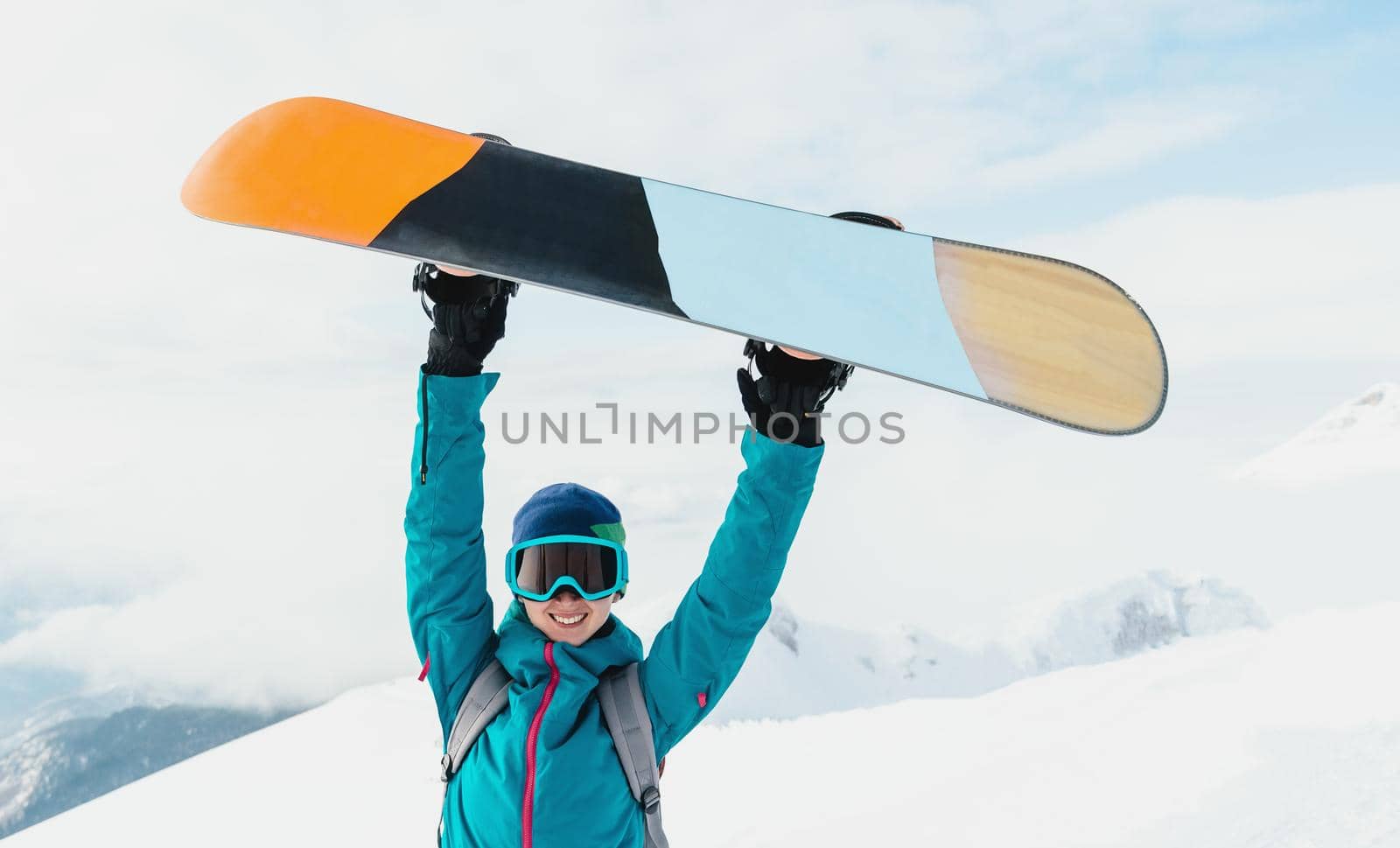 Happy girl holding snowboard over her head on snowy mountains in winter, looking at camera