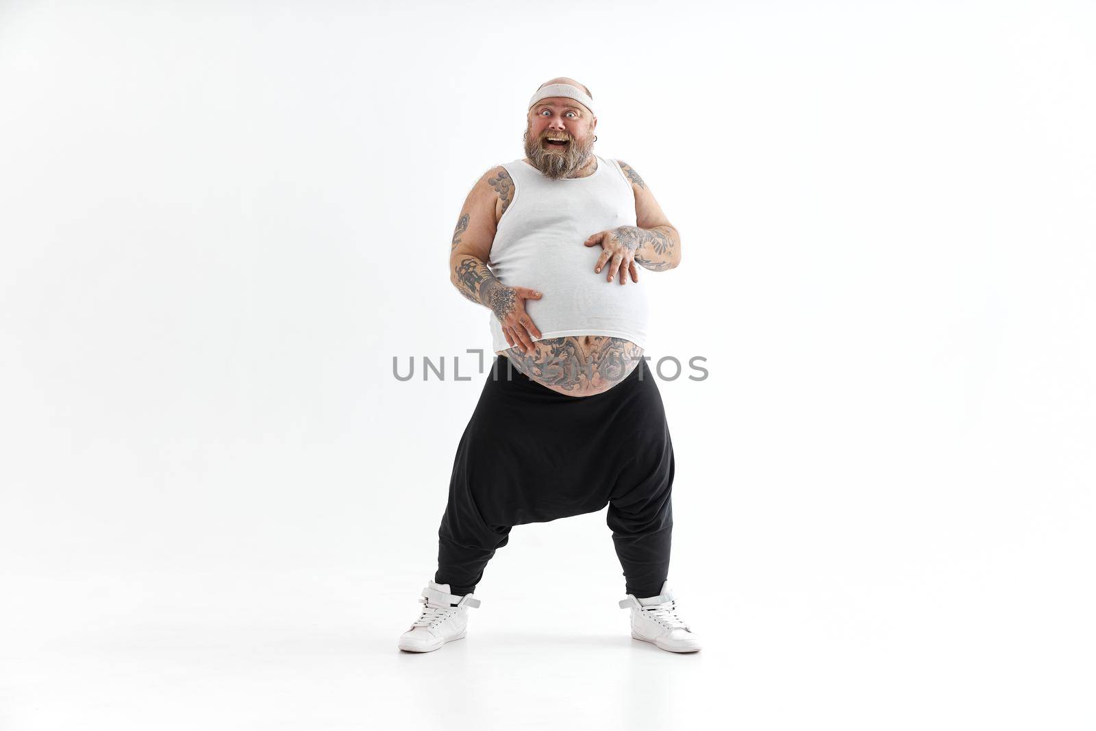 Happy fat man with big belly and tattoes in sports wear is holding his stomach