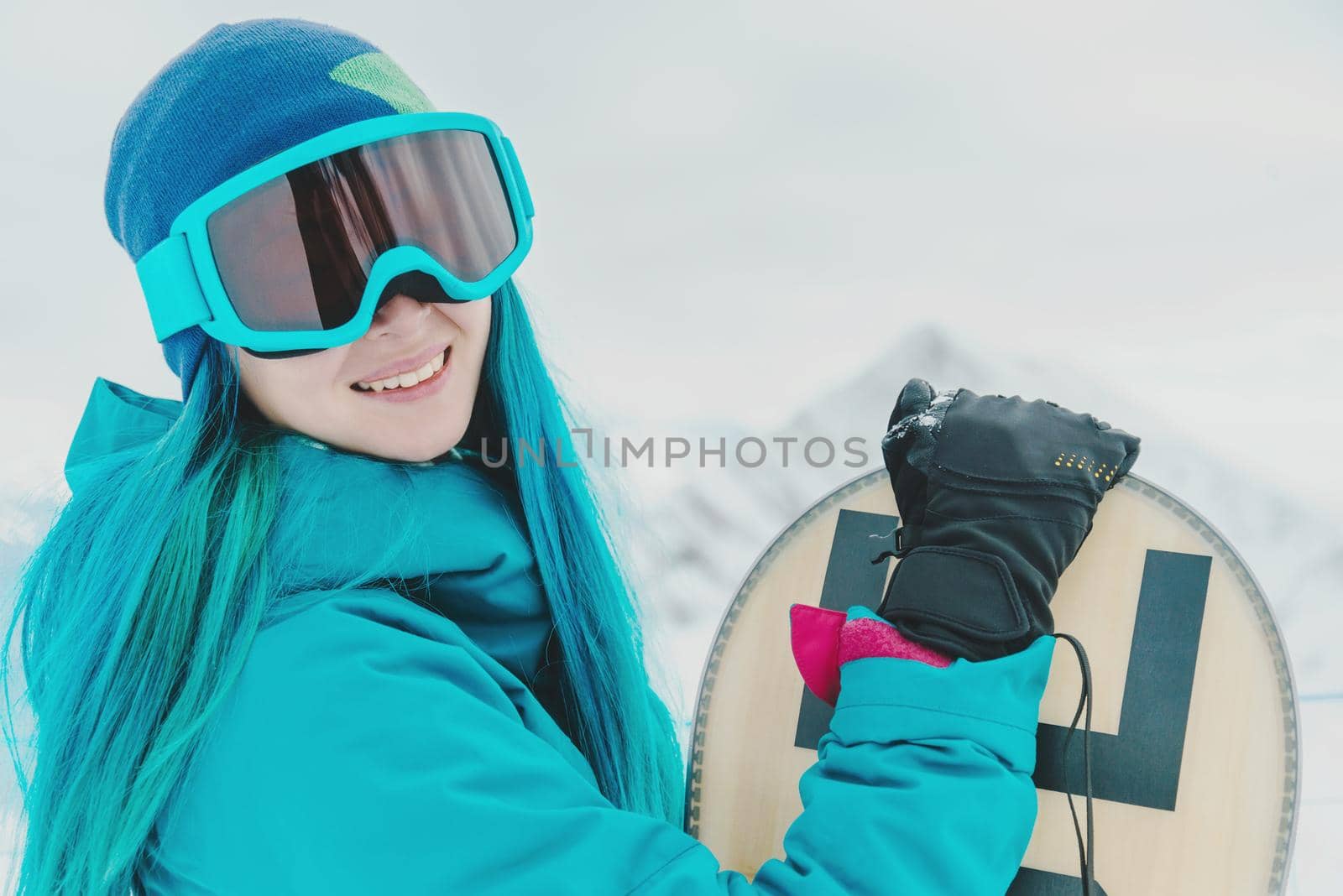 Smiling young woman with blue hair in protective sunglasses standing with snowboard on background of snowy mountains, looking at camera