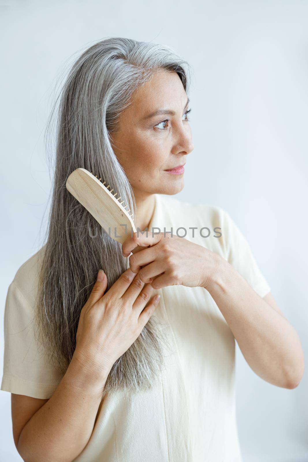 Pretty middle aged Asian person brushes straight grey hair posing on light background in studio. Mature beauty lifestyle