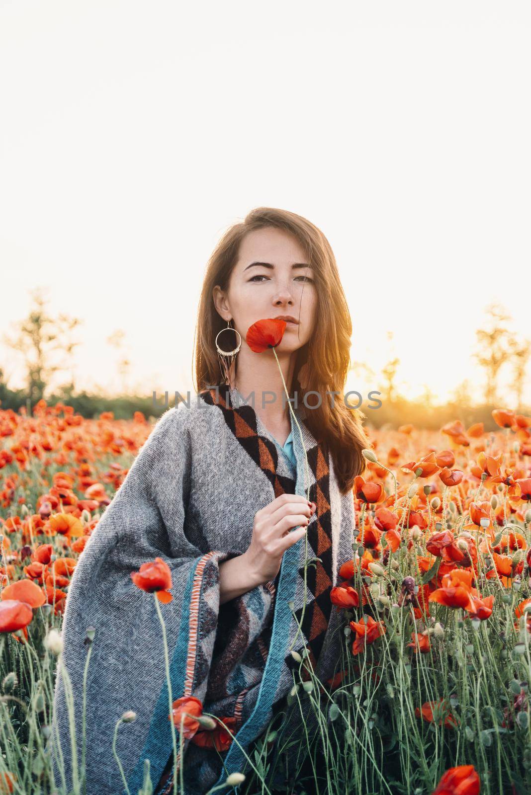 Attractive brunette young woman with long hair standing with one poppy in flower field at sunset in summer outdoor.
