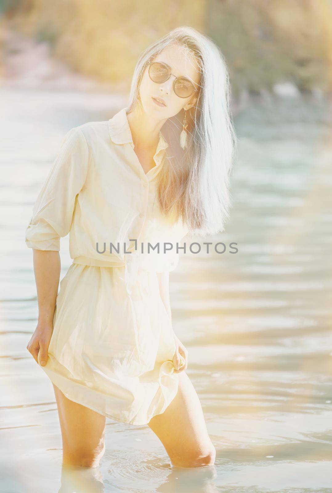 Attractive young woman with blue hair wearing in dress and sunglasses resting on lake, looking at camera. Image with sunlight effect.