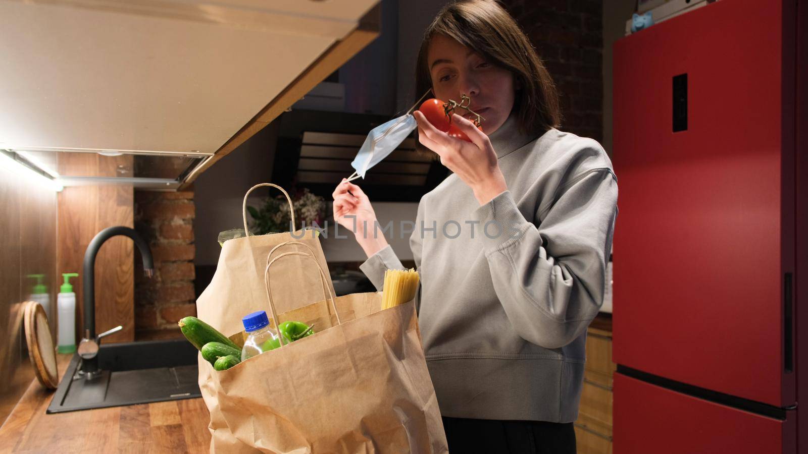 Pretty brunette woman takes off protective mask to smell fresh vegetables from large paper bags on countertop in kitchen