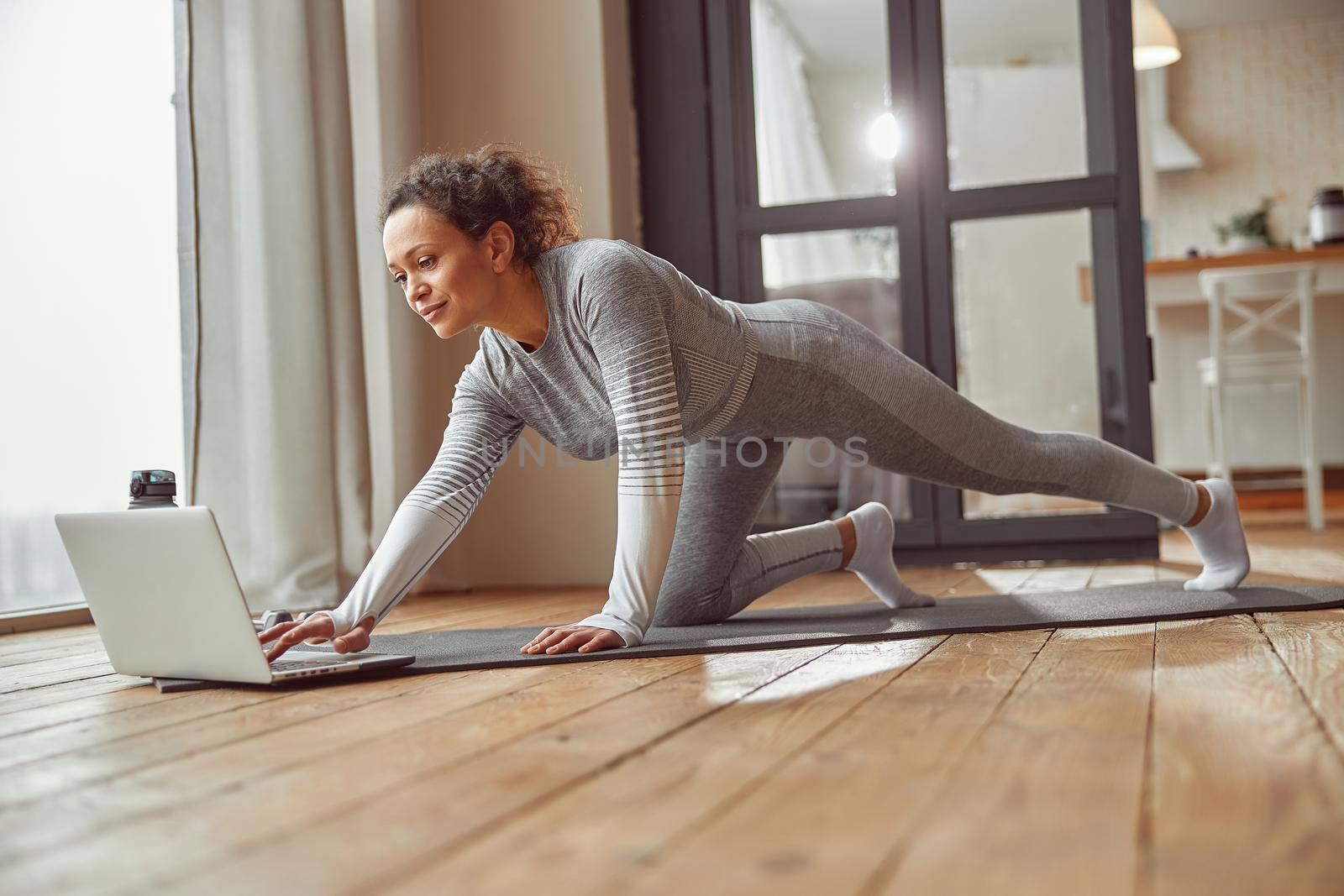 Low angle of smiling female in sportswear doing training on mat with video guide on laptop