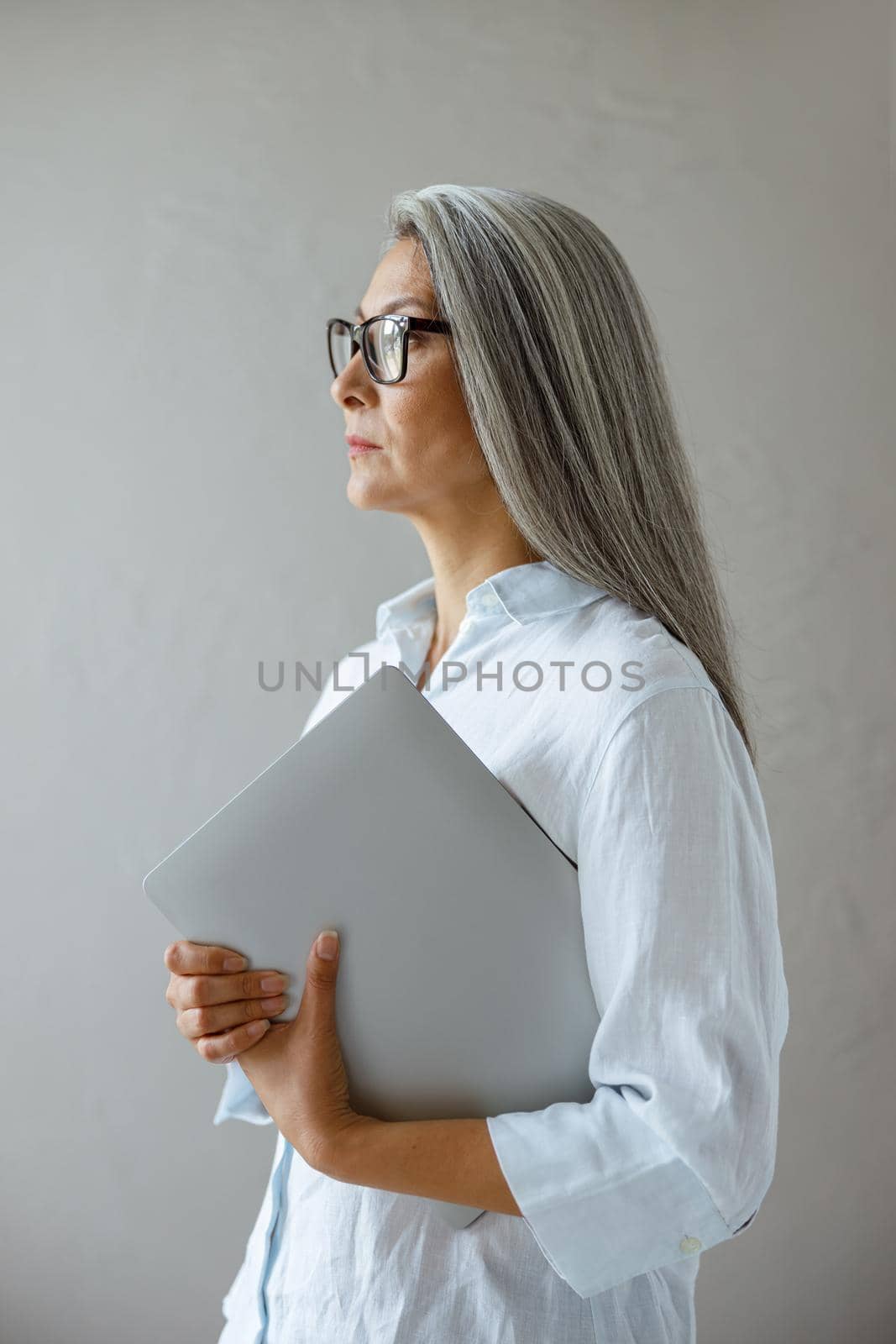 Serious mature Asian businesswoman wearing elegant blouse holds contemporary laptop standing near grey stone wall in studio side view