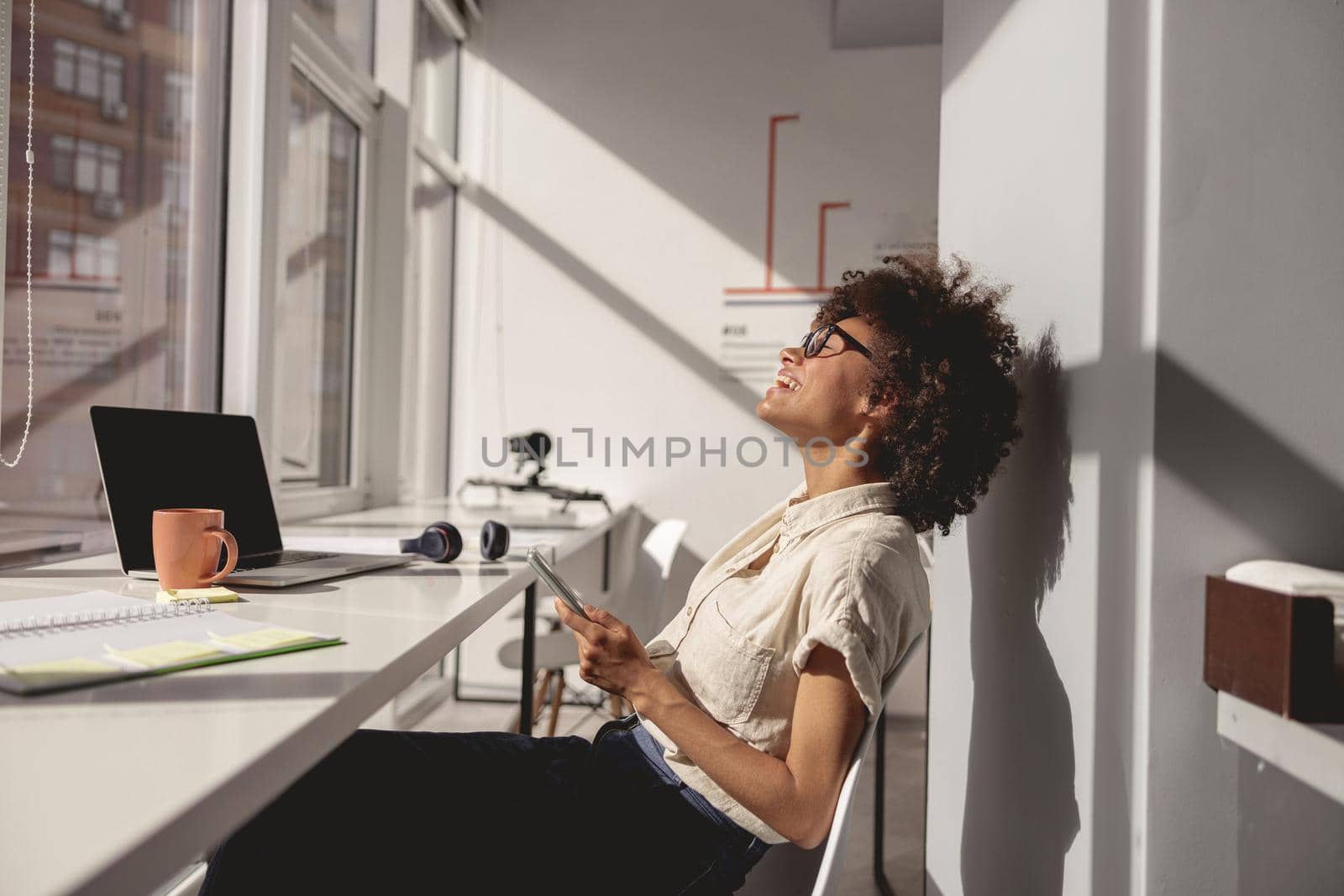 Side view of happy Afro American lady using mobile phone while working in the office near the window