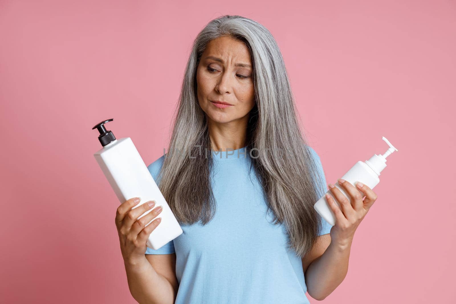 Thoughtful middle aged Asian lady chooses cosmetic product on pink background by Yaroslav_astakhov