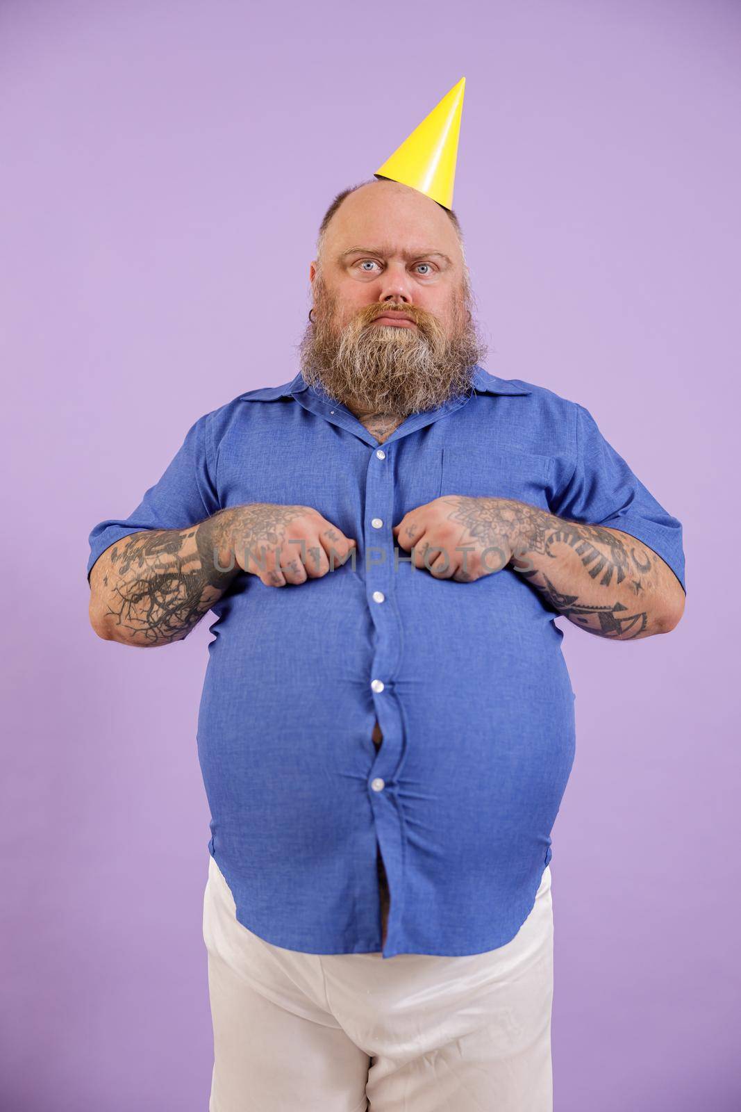 Displeasured man with overweight in party hat poses on purple background by Yaroslav_astakhov