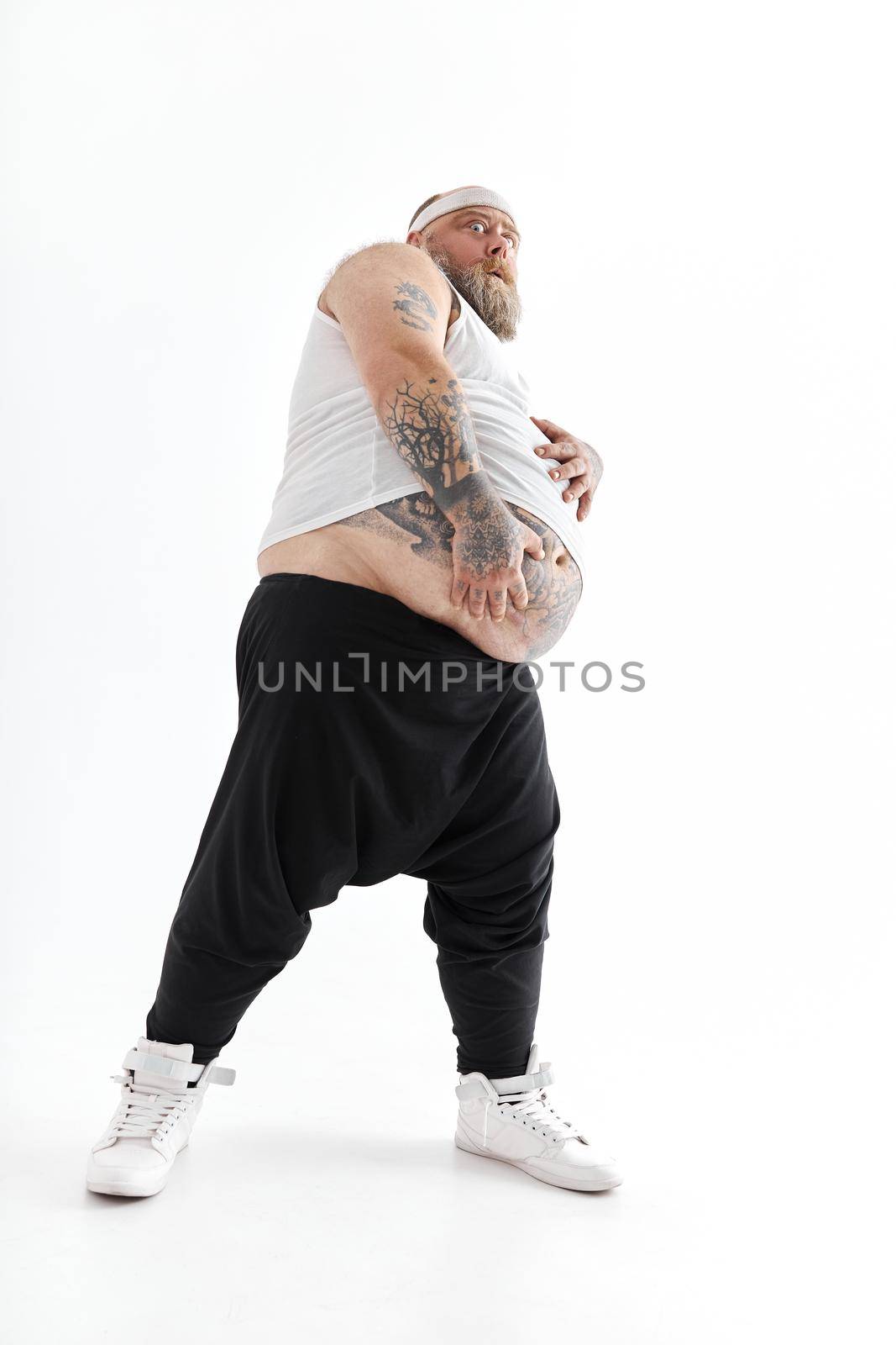 Happy fat man with big belly and tattoes in sports wear is holding his stomach by Yaroslav_astakhov